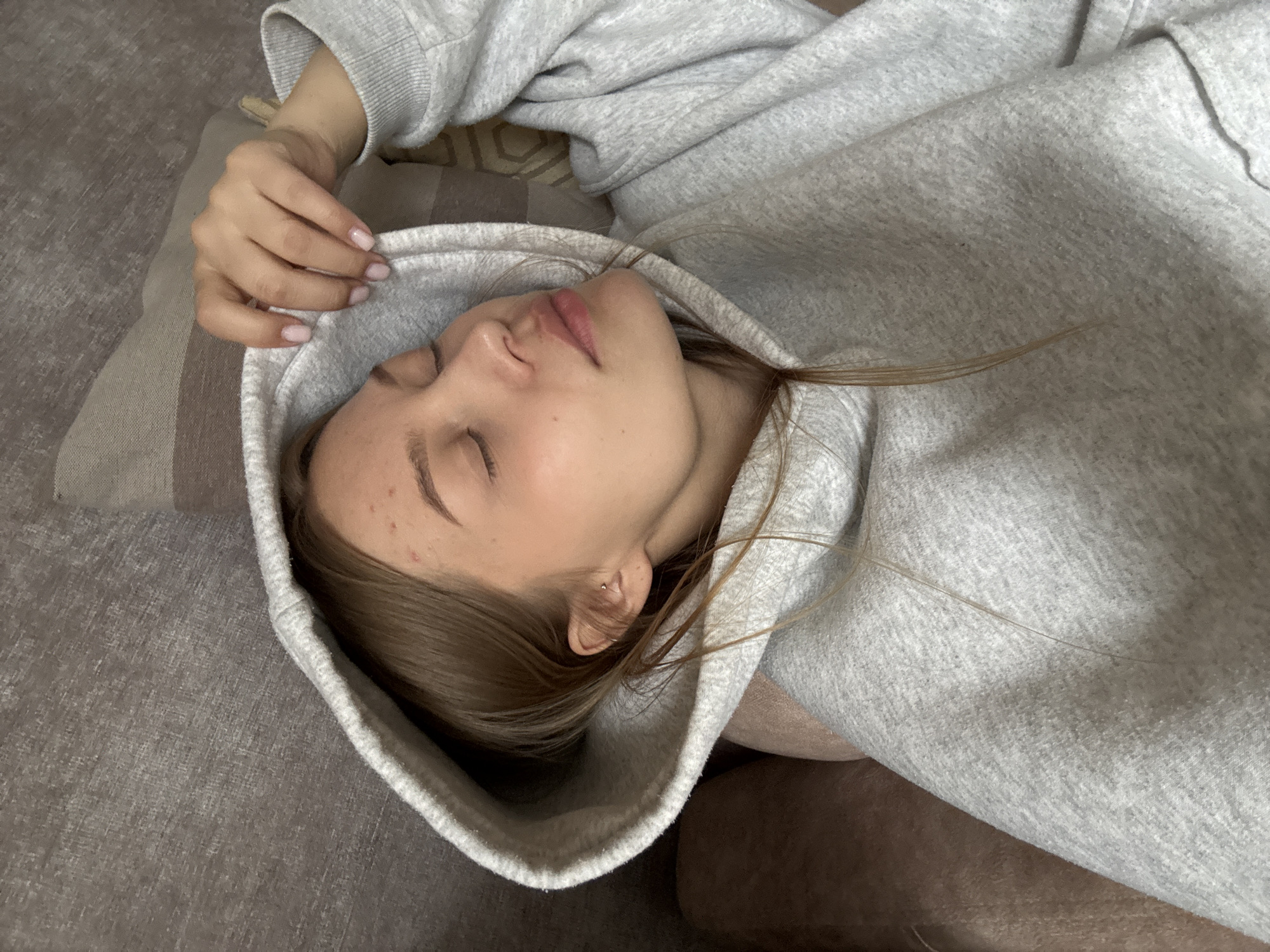 An overhead shot captures a young woman resting indoors in Chaikovsky, Russia. She is lying on her side, facing right, with her eyes closed, suggesting she is either sleeping or deeply relaxed. Her light brown hair spills slightly from beneath the hood of a light gray, possibly fleecy, hooded sweatshirt she is wearing, which frames her face.
Her complexion is fair, with some minor blemishes visible on her forehead near her hairline. Her lips are naturally pink and slightly parted. A small, simple earring can be seen in her left earlobe. Her left hand is partially visible near her head, resting on the fabric of her hoodie, showing nails painted with a light, neutral pink polish.
She is positioned on a surface that appears to be a couch or bed, covered in a textured, possibly woven, gray fabric. Behind her head and shoulder, a striped pillow or cushion in shades of brown and beige is visible. The lighting is soft and ambient, indicative of an indoor setting, with no harsh shadows to suggest a specific time of day beyond daylight or well-lit evening. No specific weather conditions are discernible. There is no visible text in the image. The overall mood is one of peaceful repose.