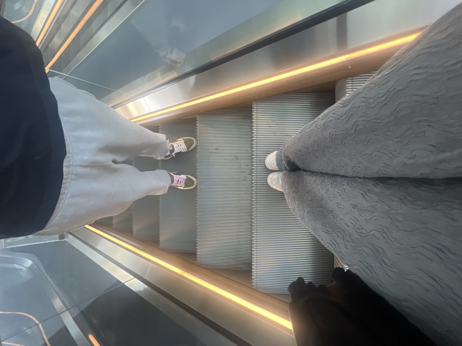 The image provides a high-angle view looking down an escalator in an indoor, modern architectural setting within the city of León, Mexico. Two individuals are visible. In the immediate foreground, the lower torso and legs of a person are shown, wearing light grey textured wide-leg pants and light-colored sneakers; a dark object is partially visible on the far left. One step below, another individual's lower torso and legs are visible, dressed in light grey sweatpants and two-toned sneakers, one with pink laces and the other with white or cream laces, both featuring green/earth-toned bases. The escalator steps are metallic and ribbed. Transparent glass balustrades flank the steps, with metallic handrails above them. A warm orange-yellow LED light strip runs along the base of the handrail on both sides of the escalator. Both subjects are standing stationary on the moving escalator.