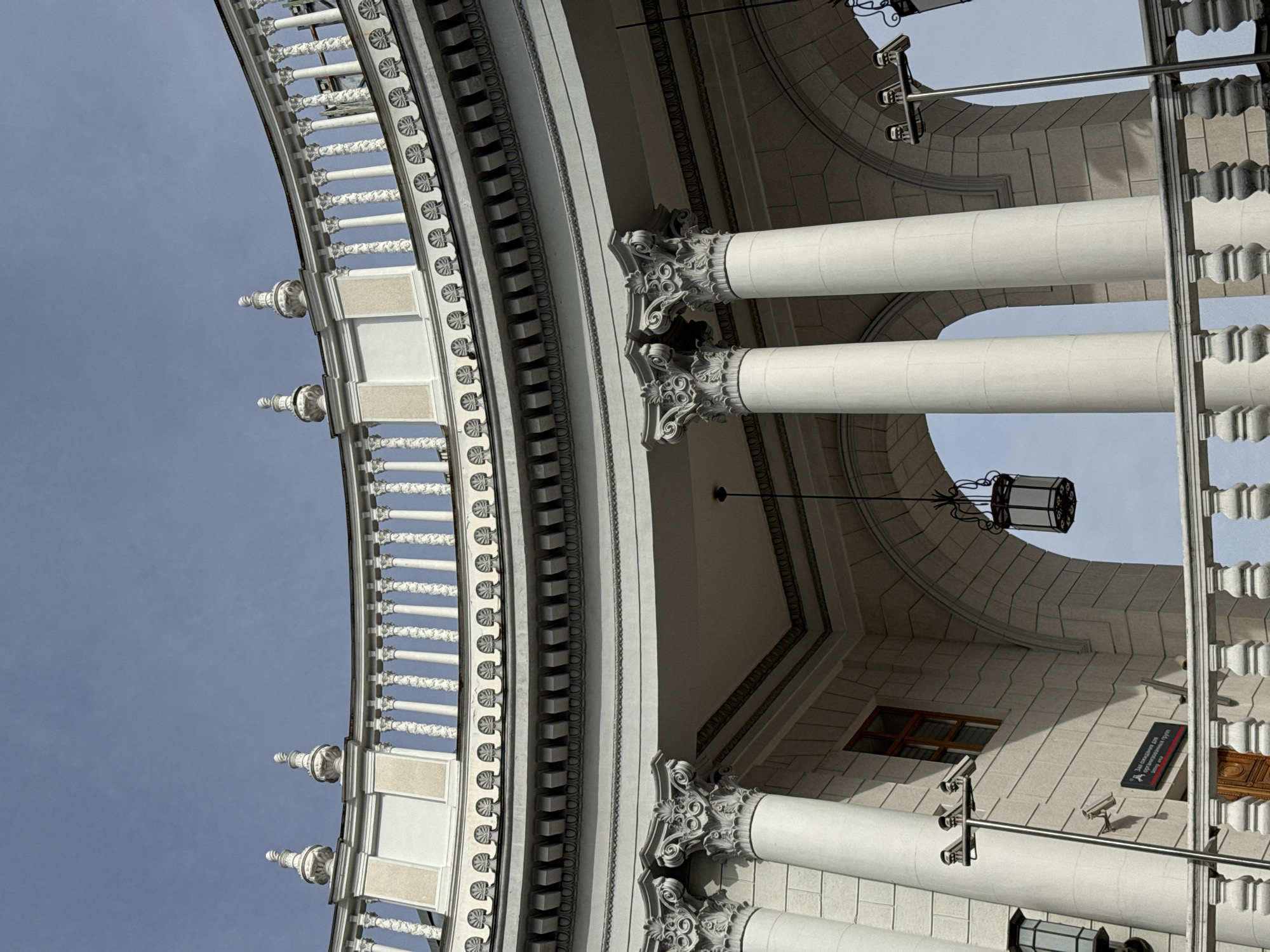 The image captures an upward view of an ornate, neoclassical architectural structure. Prominent features include rows of white columns topped with elaborate, floral capitals and detailed balustrades with decorative elements. A portion of the building's façade is visible, constructed of stone with a grid pattern, and a few security cameras are mounted on the wall. An old style lamppost hangs near the arch of the roof. The sky is visible in the background, suggesting an outdoor location. The building is located in the city of Sochi, Russia.