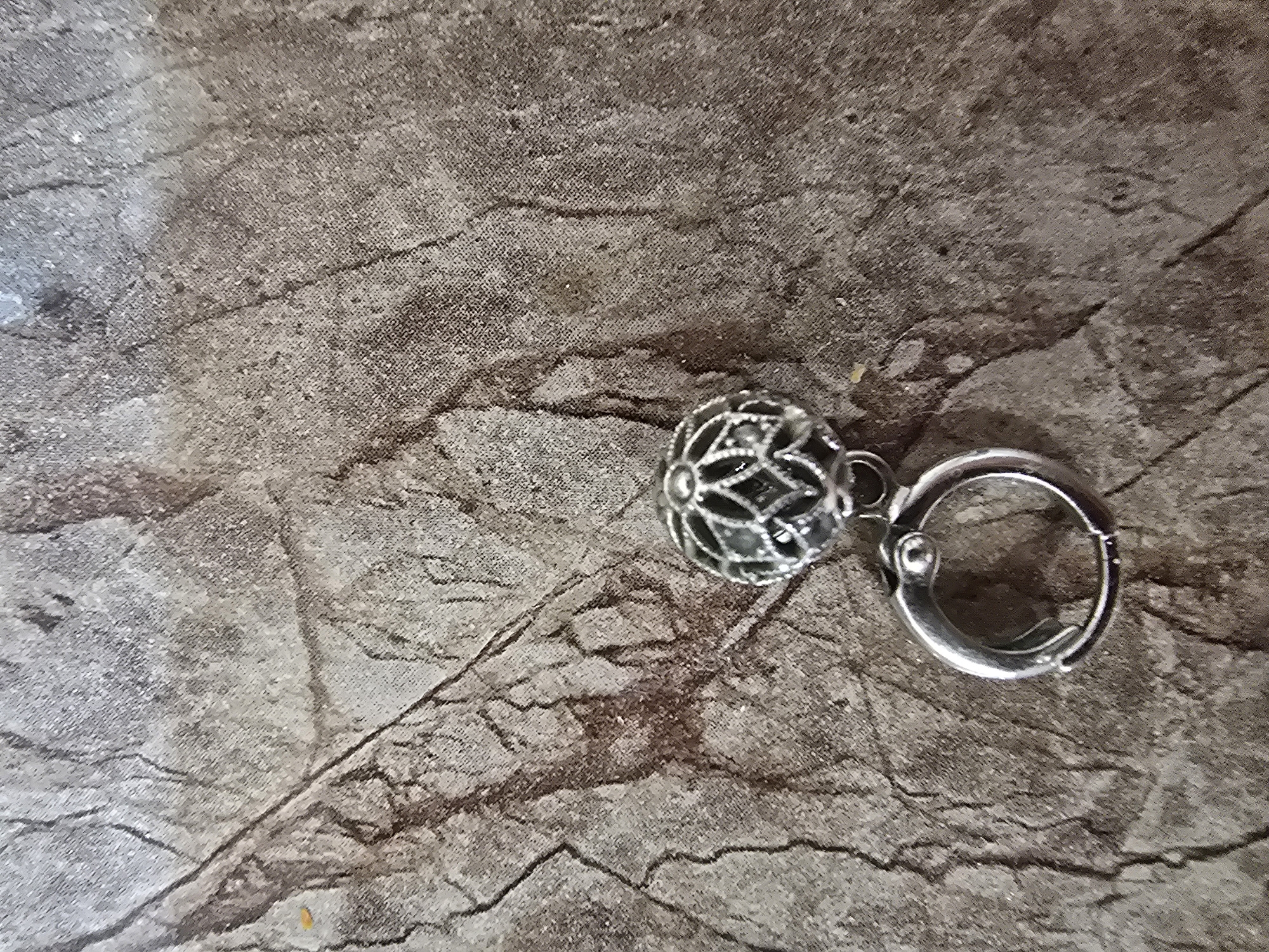 The image is a close-up, high-angle shot of a silver-colored charm with an intricate filigree design, attached to a small silver clasp ring. The charm is spherical and features a repeating pattern of diamond shapes and floral motifs. It hangs from the ring by a small connector. The charm and ring are positioned towards the bottom center of the frame. The background is a textured surface that resembles weathered wood or stone, with a pattern of dark, irregular lines and lighter, mottled areas. There is a slight yellowish speck on the background to the left of the charm. The lighting is diffused, casting minimal shadows. The image does not contain any identifiable landmarks or clues to suggest its location in Ulan-Ude, Russia.