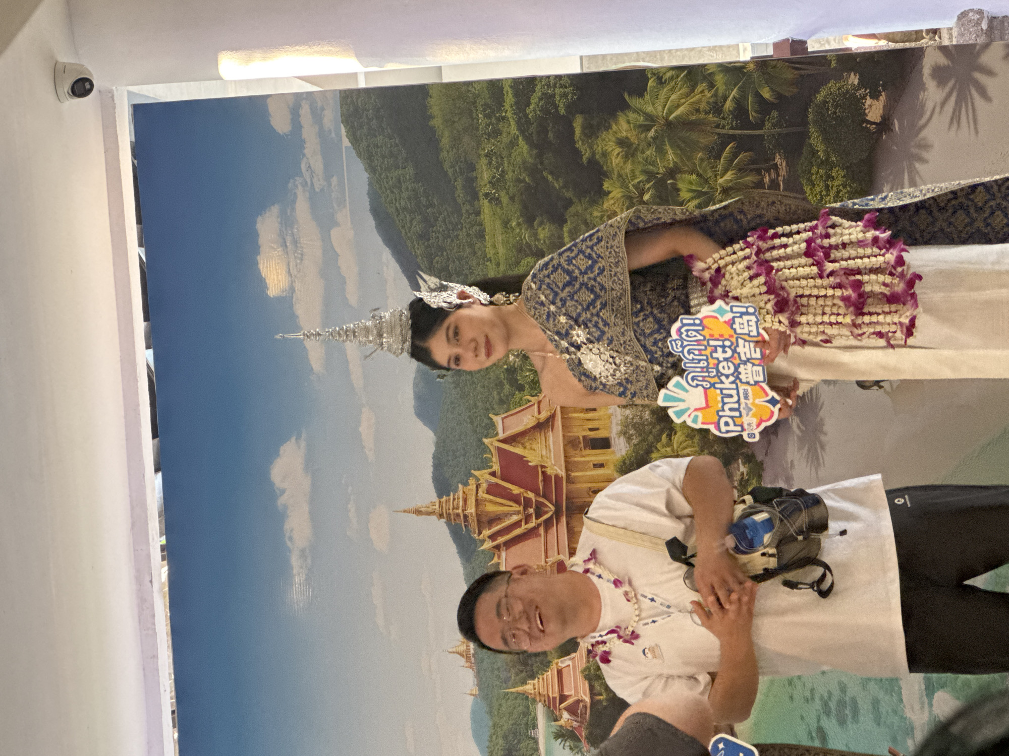 An indoor photograph captures a male tourist posing for a picture next to a woman in traditional Thai attire, against a large scenic backdrop. The backdrop depicts a vibrant Thai landscape, featuring lush green mountains, palm trees, a clear blue sky with some white clouds, and traditional gold-colored temple architecture (wats), suggesting a coastal region like Phuket.
On the left side of the image, a woman, likely a greeter or performer, is dressed in an ornate traditional Thai costume. She wears a blue and gold patterned top, a light-colored lower garment, and an elaborate silver pointed headdress. Around her neck and draped over her arm are garlands of white and purple flowers, resembling jasmine and orchids. She holds a small, colorful sign that prominently displays "สวัสดี Phuket!" (Hello Phuket!) and "Phuket!" in English, along with Chinese characters for "Welcome." She smiles faintly while looking towards the camera.
On the right, a man with short dark hair and glasses, wearing a white t-shirt, stands smiling widely at the camera. He has a floral garland around his neck, similar to the one worn by the woman, and clasps his hands together in front of him, potentially in a gesture of greeting or excitement. A dark fanny pack or small shoulder bag is slung across his chest.
A white dome security camera is mounted on the wall in the upper left corner, and a strip of artificial lighting is visible on the ceiling. The overall scene captures a lively tourist moment, likely at an entry point or attraction in Patong, Thailand, designed to welcome visitors. The bright lighting and joyful expressions contribute to a cheerful atmosphere.