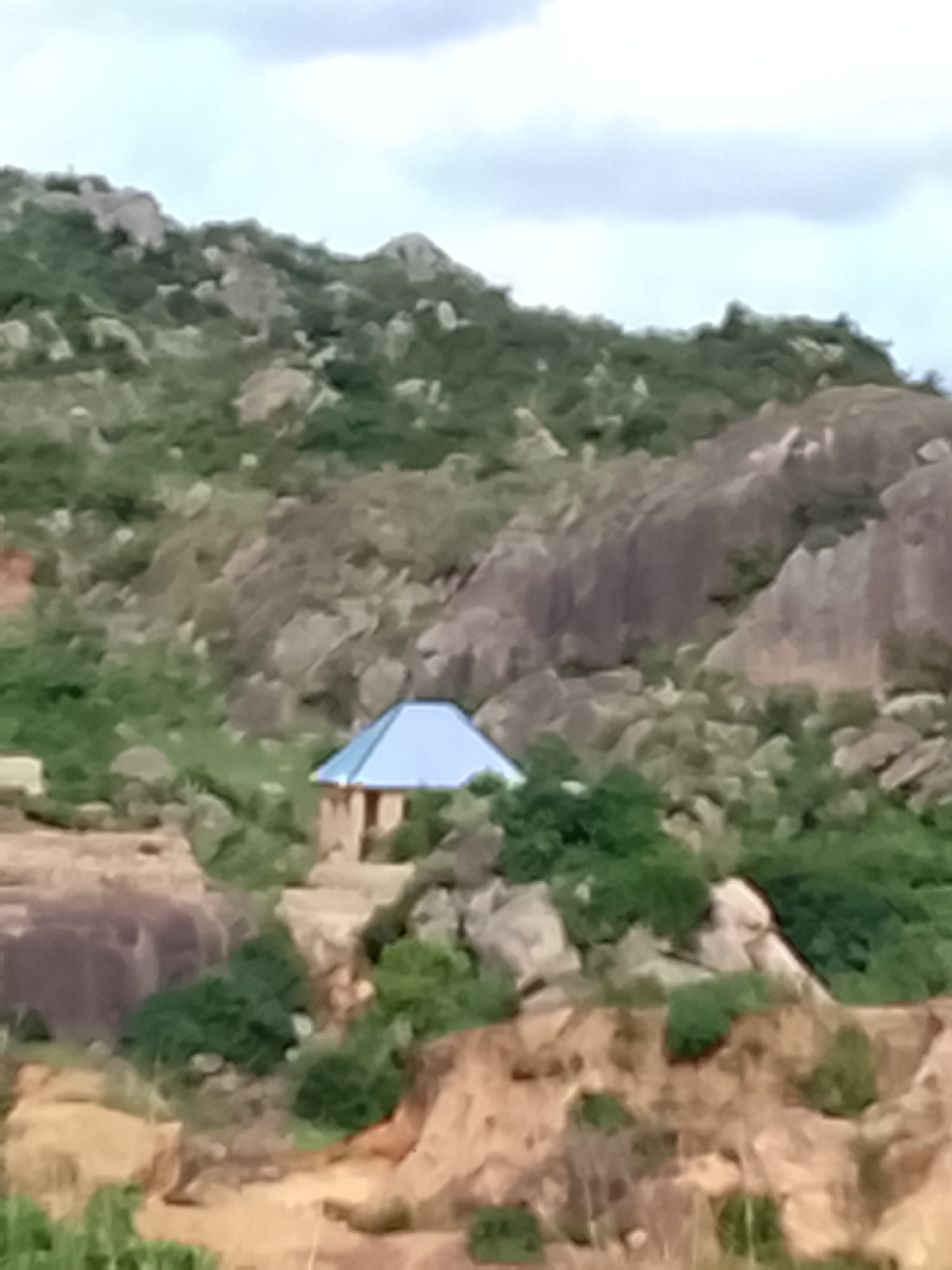 A small, tan-colored structure with a light blue, pitched roof is situated among rocky hills and green vegetation in Rafin Bauna, Nigeria. The structure appears to be a single room with visible doorways. The surrounding environment is characterized by large, weathered rocks, scrubby bushes, and a cloudy sky. The ground in the foreground consists of sandy, eroded earth with sparse green growth. The overall scene depicts a rural or semi-rural landscape.