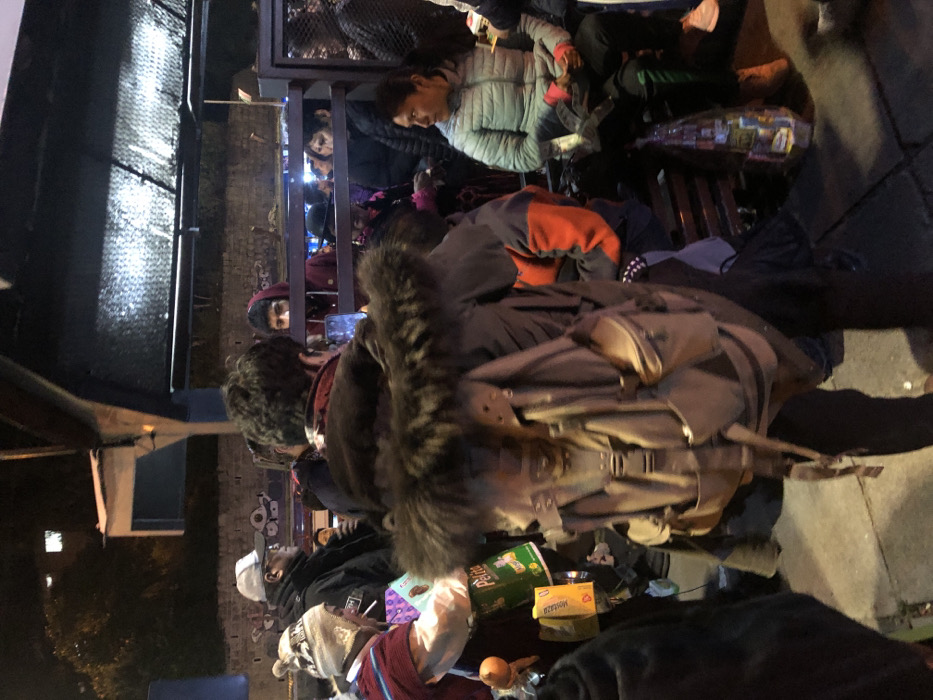 People sit on wooden platforms bundled in coats, hats, and scarves. A backpack with a fur-lined hood rests in the foreground. Snacks, including a box of mustard and a green bag of chips, are scattered on the ground. Some people are looking up, possibly at an unseen event. A phone is being held up to capture the moment.
