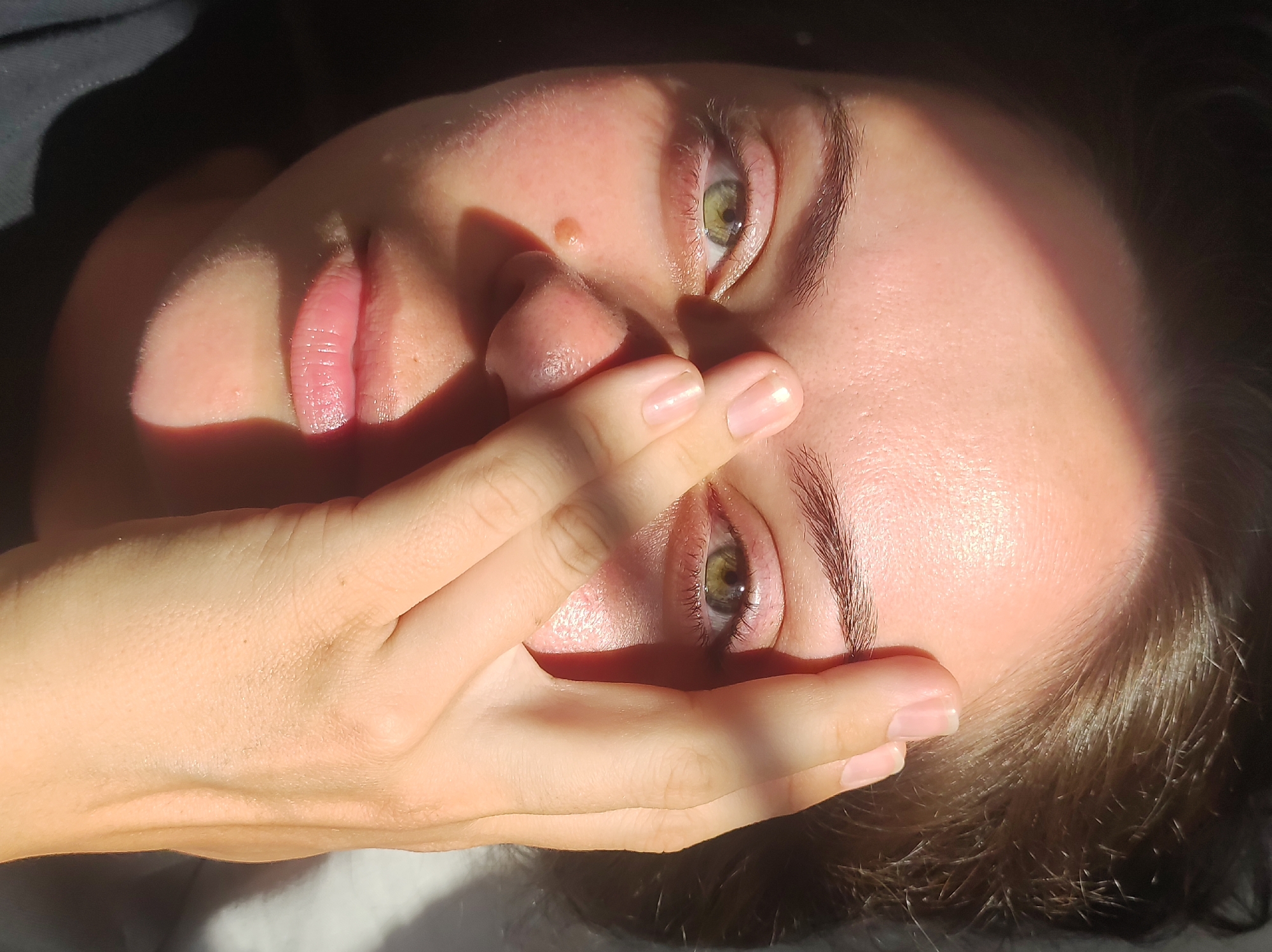 The image is a close-up, angled shot of a person's face. The person is lying down, with their head tilted. Their eyes are green and their lips are pink. A hand is covering part of their face, with fingers partially obscuring their eyes and nose. Sunlight is casting shadows across the face, highlighting the contours and textures of the skin. The background is dark and out of focus, with some lighter fabric visible at the bottom. There is no indication of the location in Chaikovsky, Russia.