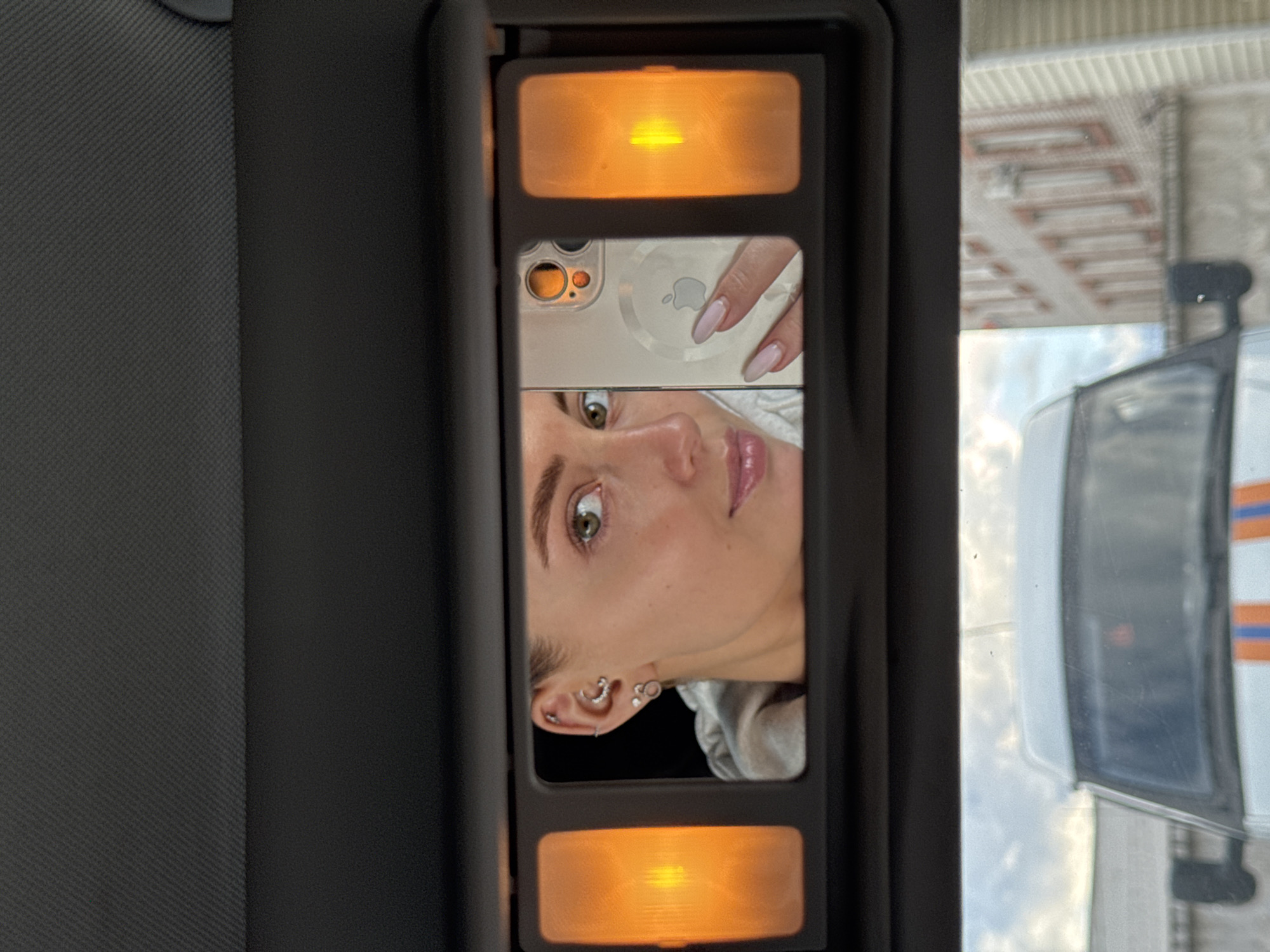 The image, taken inside a vehicle, focuses on a vanity mirror attached to a sun visor. The mirror reflects a young woman's face, a hand holding a smartphone, and the smartphone itself.
In the center of the mirror, the woman's face is visible, with her green eye looking slightly away from the camera. Her lips are slightly parted and appear glossy. Her left ear, adorned with multiple piercings including a helix, conch, and lobe piercings, is prominent. Above her face, her hand with light pink/nude manicured nails holds a smartphone. The phone case is light-colored, possibly clear, showing an Apple logo and multiple camera lenses.
The vanity mirror is flanked by two warm, amber-toned lights, one at the top and one at the bottom, which are illuminated. The mirror and lights are set into a dark, rectangular panel. To the left, the dark gray, textured fabric of the car's interior (likely the headliner or visor) is visible.
Outside the car window, to the right of the mirror, an exterior scene is visible. Parts of a building with horizontal structures are discernible. A white vehicle with orange and blue stripes, possibly an emergency or utility vehicle, is reflected, along with a black T-shaped object on its roof. The sky appears overcast. The overall lighting inside the car is a mix of the warm vanity lights and the cooler daylight from outside.