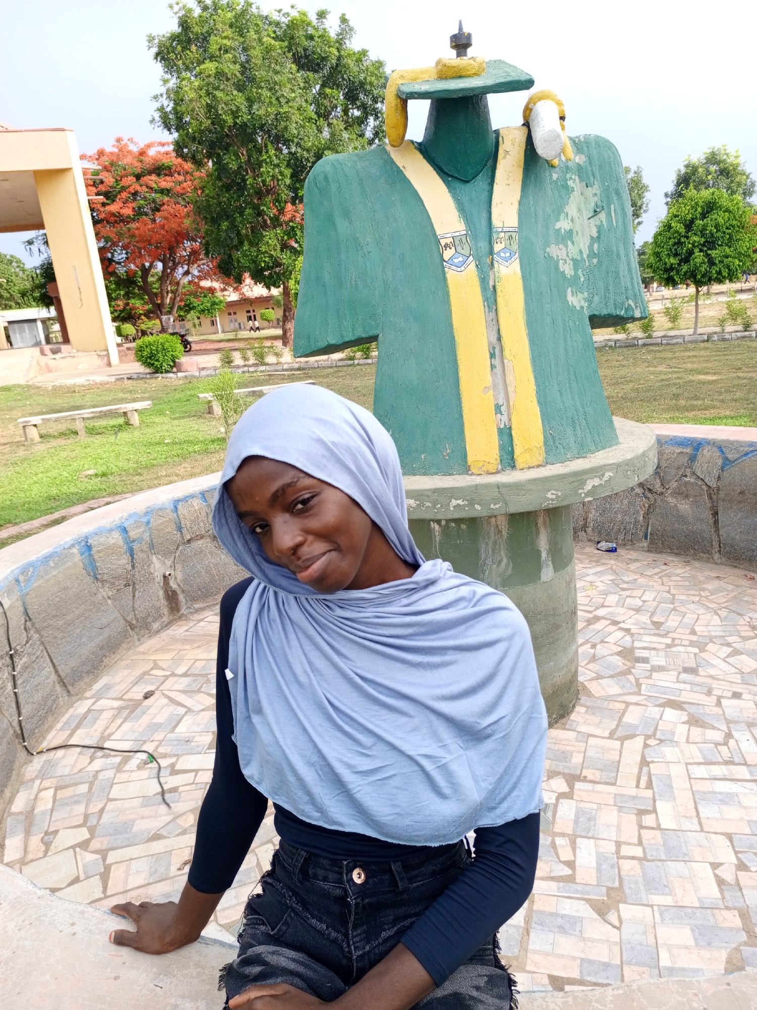 The image features a young woman wearing a light blue headscarf, a black long-sleeved shirt, and dark denim shorts. She is sitting on the edge of a low, decorative wall made of patterned stone tiles. Behind her stands a large, weathered statue of a graduation gown with a mortarboard hat, painted in green and yellow.
The setting appears to be a public park or campus area. In the background, there is a grassy lawn, trees, and a building with a pale yellow exterior. A red-flowered tree is visible on the left. The location is identified as Unknown, Unknown.