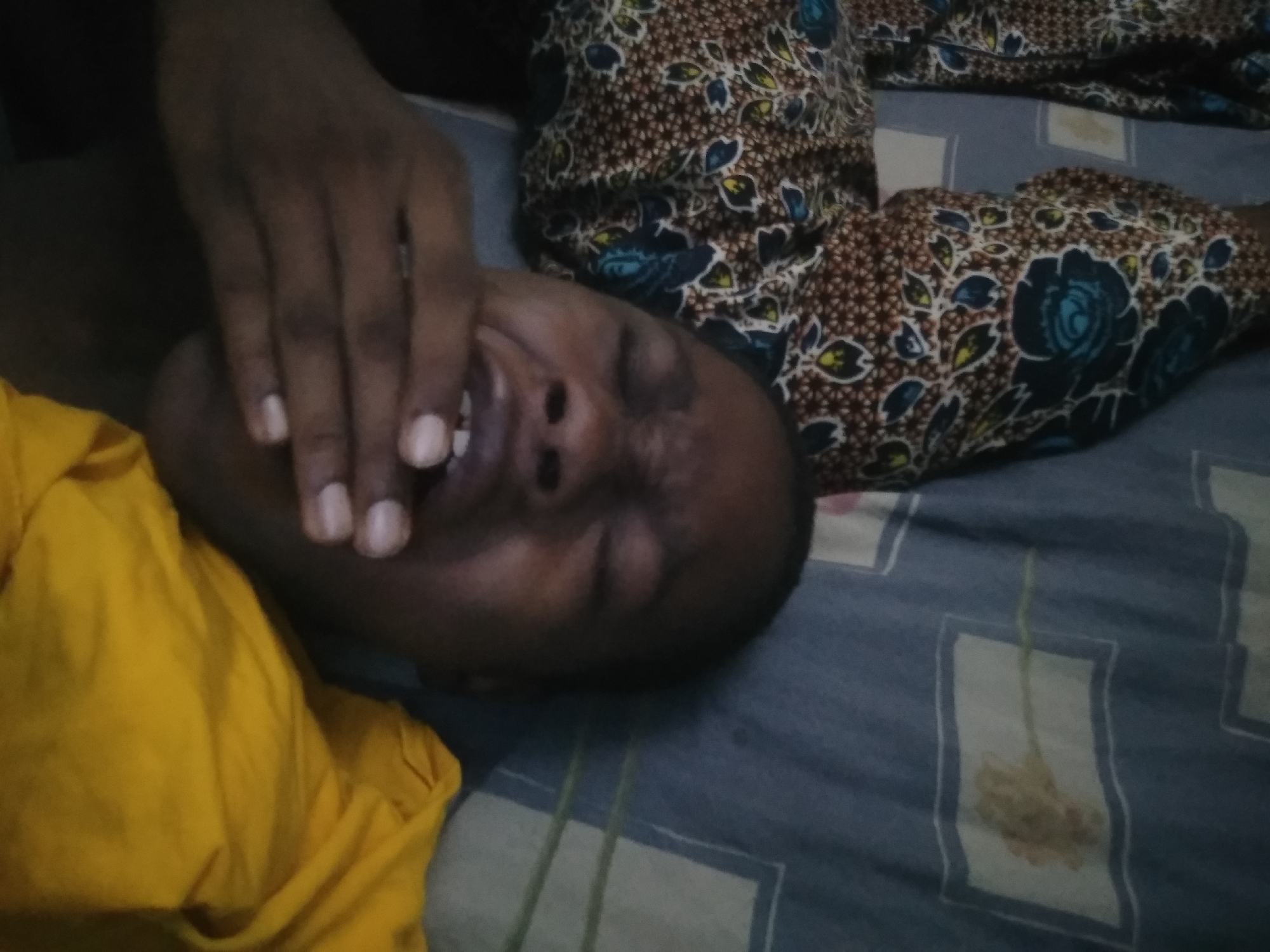 A person is lying on a bed with a hand covering their mouth. The person is wearing a yellow shirt and is lying on a blue and white patterned bedsheet. A patterned cloth is visible next to the person. The location is Shangisha, Nigeria.