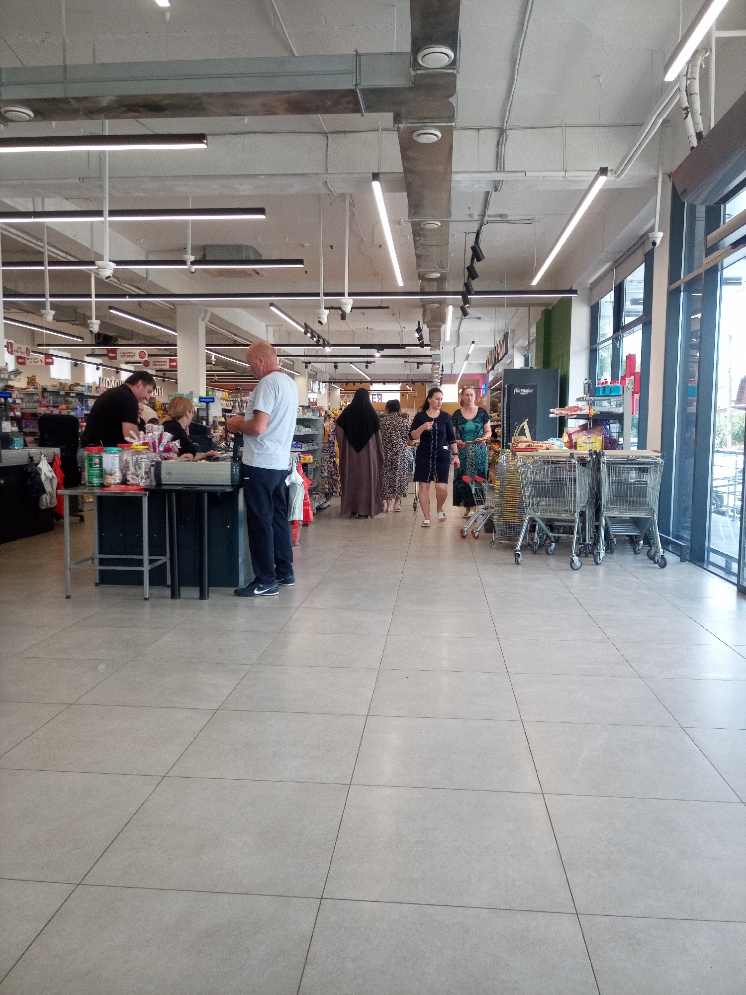 This is an eye-level, full shot of the interior of a supermarket in Nalchik, Russia. In the foreground, a bald man in a gray t-shirt and dark pants stands at a checkout counter, facing away from the camera, interacting with a cashier. To his left, a woman in a black top and light-colored pants is also at the counter, looking towards the cashier. Behind them, another cashier is visible. The checkout area is equipped with a scale and various items on display. Further back, several shopping carts are neatly arranged. To the right, two women are walking through the aisle, one in a dark blue dress and the other in a green patterned dress. In the background, shelves stocked with goods line the walls, and a person wearing a dark veil and a long, flowing brown garment is visible at the far end of the aisle. The floor is tiled with large, light gray squares. The environment is brightly lit with fluorescent ceiling lights.