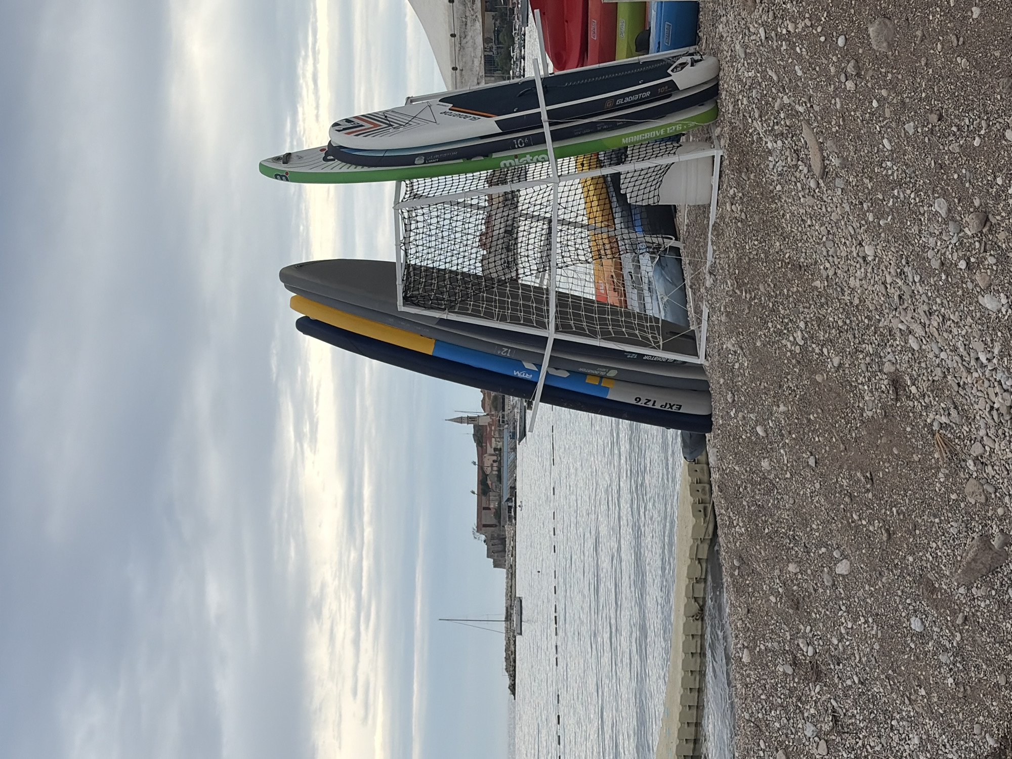 The image shows a rack of paddleboards and kayaks on a pebble beach next to calm water, with a view of the city of Budva, Montenegro, in the background. The rack is white and metallic, with netting on some of the levels. Several paddleboards are stacked on top, including models labeled "GLADIATOR 10'6", "MANGROVE 12'6", and "EXP 12.6". Kayaks are also visible. A small boat floats on the water, and the skyline of Budva is visible in the distance, featuring buildings and a church spire. The sky is overcast. The beach is made of light-colored pebbles and sand. A low, gray retaining wall separates the beach from the water.