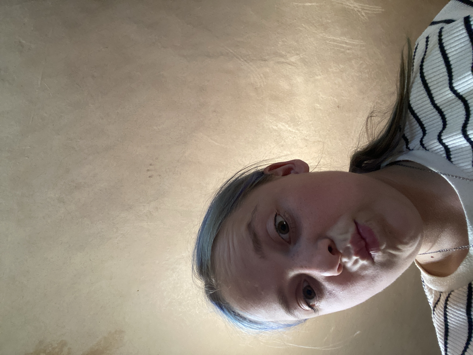 The media file is a close-up, downward-facing selfie of a young person in Nova Praha, Ukraine. The subject has fair skin, brown eyes, and blue-tinted hair. They are wearing a white and black striped sweater and a silver necklace. The person's mouth is slightly open, and they appear to be making a playful or curious expression. The background is a textured, light-brown surface, possibly a wall or ceiling, with a subtle sheen. There are no clear indicators of the time of day, weather, or specific location beyond the provided context of Nova Praha. The overall mood is informal and candid.