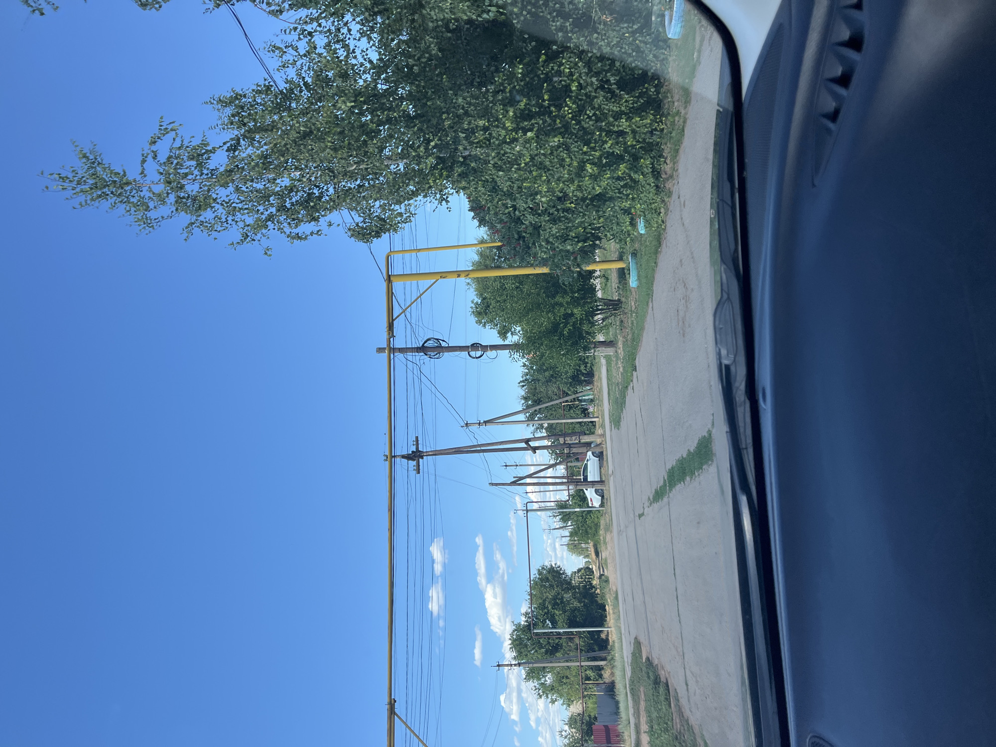 This image depicts a roadside scene in Rakhinka, Russia, on a clear day. The perspective is from inside a vehicle, likely a car, looking out the side window.
The primary subjects are a paved road running alongside a grassy area, with numerous utility poles and overhead wires stretching across the frame. Dense green trees and foliage line the road, some of which overhang the roadway. In the distance, a white car is visible. The sky is a vibrant, cloudless blue, suggesting fair weather and likely daytime. A yellow structure, possibly part of an electrical substation or a similar piece of infrastructure, is prominent in the mid-ground. The interior of the car, specifically the dark dashboard and part of the door panel, is visible on the right side of the image, indicating the viewpoint. The overall scene suggests a rural or semi-rural setting. No people are visible, and there is no visible text.