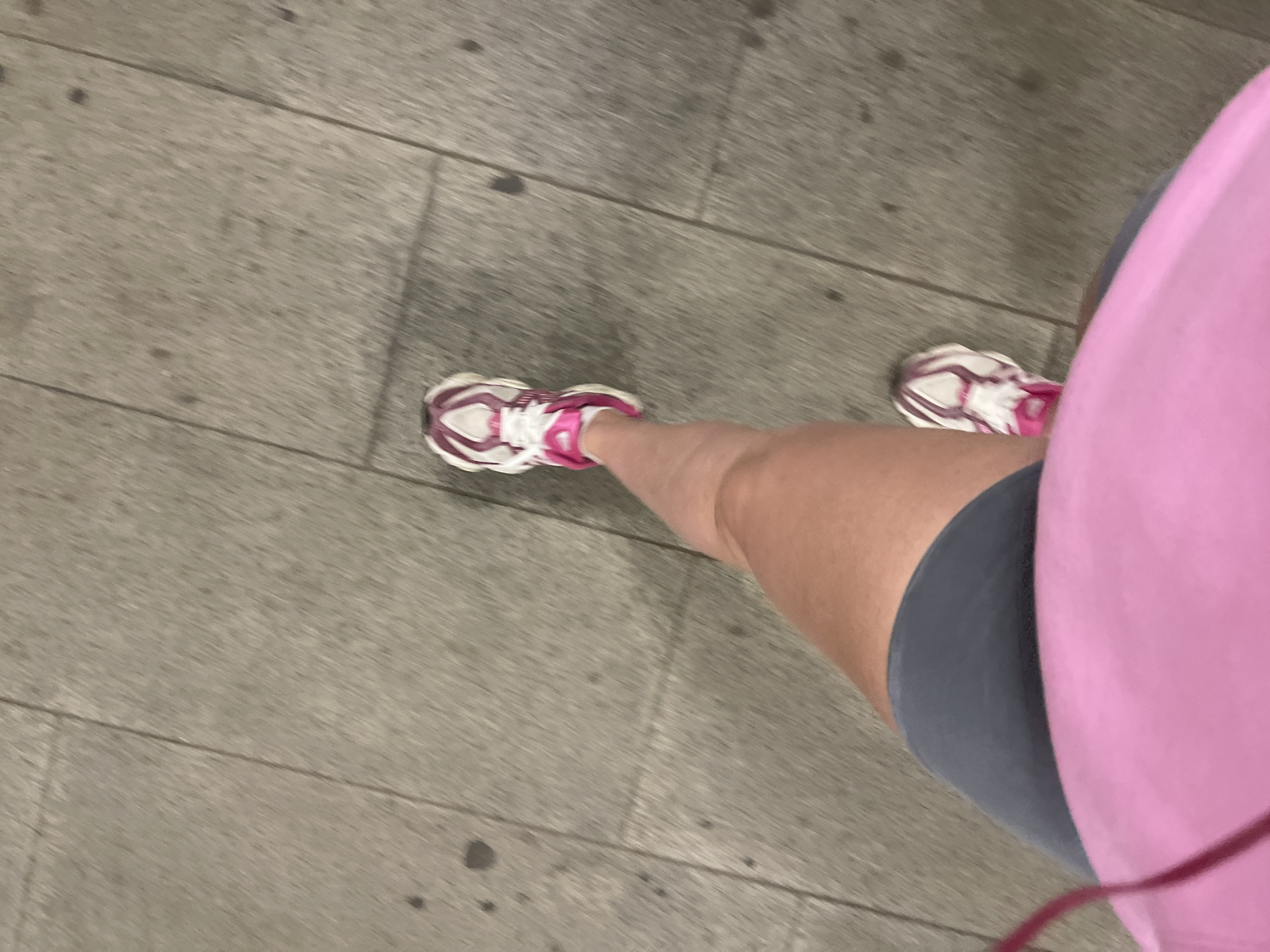 The image shows a person's legs from a first-person perspective, standing on a paved surface. The person is wearing pink and white athletic shoes, gray shorts, and a pink top. The background is a gray paved surface. The location is unknown.