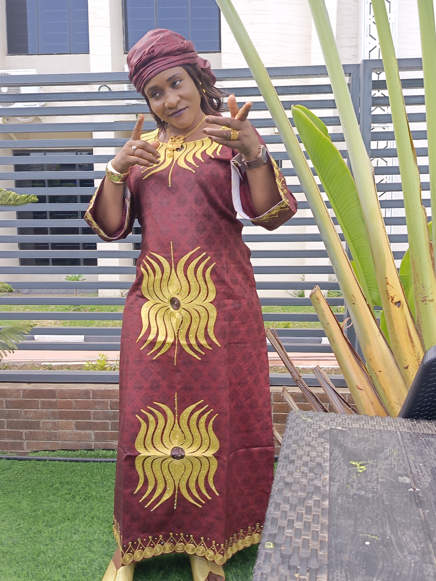 A woman is captured in a full-shot outdoor portrait, posing in what appears to be a garden or courtyard in Akwanga, Nigeria. She is the central subject, wearing a long, loose-fitting reddish-brown dress, possibly a kaftan, adorned with striking gold embroidery. A prominent gold floral or abstract design is featured on her torso, and a detailed gold patterned border decorates the bottom hem of her dress. She also wears a matching reddish-brown headwrap.
Her accessories include a gold necklace, gold rings on both hands, a gold-colored bangle on her left wrist, and a watch on her left wrist. Gold-toned footwear is partially visible at the bottom. The woman has both hands raised, with her index fingers pointing outwards in a confident and possibly playful gesture. She looks slightly to her right with a faint smile on her face.
The background consists of a modern building with large dark windows, partially obscured by a grey horizontal slatted fence. To the right, several large, green plant stalks, possibly from a Traveler's Palm, frame the scene. The foreground features manicured green grass and a section of a brick wall. A dark, woven outdoor table is partially visible on the right side. The natural lighting indicates it is daytime. There is no visible text in the image.