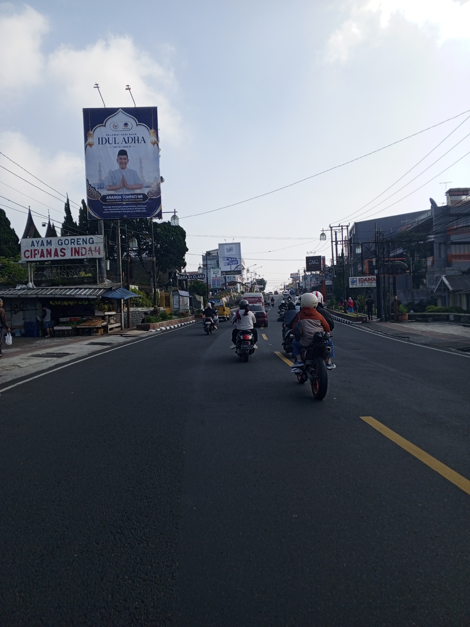 This image depicts a street scene in the city of Weddagala, Sri Lanka, captured during daylight hours under a bright, partly cloudy sky. The perspective is from a low angle, looking forward along a wide asphalt road marked with a solid yellow line on the right edge of the visible lane.
On the left side of the road, a large billboard is mounted on a tall pole. The billboard features the Indonesian text "SELAMAT HARI RAYA IDUL ADHA 1447 H / 2026 M" (Happy Eid al-Adha 1447 H / 2026 AD) and displays a portrait of a smiling man. Below the portrait, the text identifies him as "ANANDA TOHPATI NR, ANGGOTA DPR RI - FRAKSI PARTAI NASDEM" (Member of the House of Representatives - Nasdem Party Faction) and includes an Indonesian quote: "Semoga semangat pengorbanan, keikhlasan dan kepedulian terus tumbuh di hati kita untuk membangun bangsa yang lebih baik." (May the spirit of sacrifice, sincerity, and care continue to grow in our hearts to build a better nation). Beneath the billboard, a roadside stall with a corrugated metal roof is present, displaying a sign with Indonesian text "AYAM GORENG CIPANAS INDAH" (Fried Chicken Cipanas Indah). Produce, including bananas, is visible at the stall. Two individuals are near this stall, one partially obscured by the foreground. Coniferous trees and numerous utility lines on poles are visible in the background on the left.
On the road, multiple motorcycles are traveling in the same direction as the camera. Closest to the viewer, a black motorcycle with a red rear wheel carries two riders; the front rider wears an orange/brown jacket and a white helmet, and the passenger wears blue jeans. Ahead, another person in a dark helmet and light shirt rides a separate motorcycle. Additional motorcycles and a red car are visible further down the road.
The right side of the road features a paved sidewalk with a curb, some vegetation, and multi-story buildings. Several signs are visible on this side, including "STIEBIS 69" on a dark building, "IDAMA" on a lower sign, and a brown sign for "BIARA S. CLARA" (Convent of St. Clara). A more distant billboard displays the word "CLASSY" and numerical information "50 ribu 39,000". A group of pedestrians, some dressed in red, are walking on the right sidewalk in the mid-ground. The scene depicts an active urban or semi-urban environment with ongoing vehicle traffic and commercial establishments.