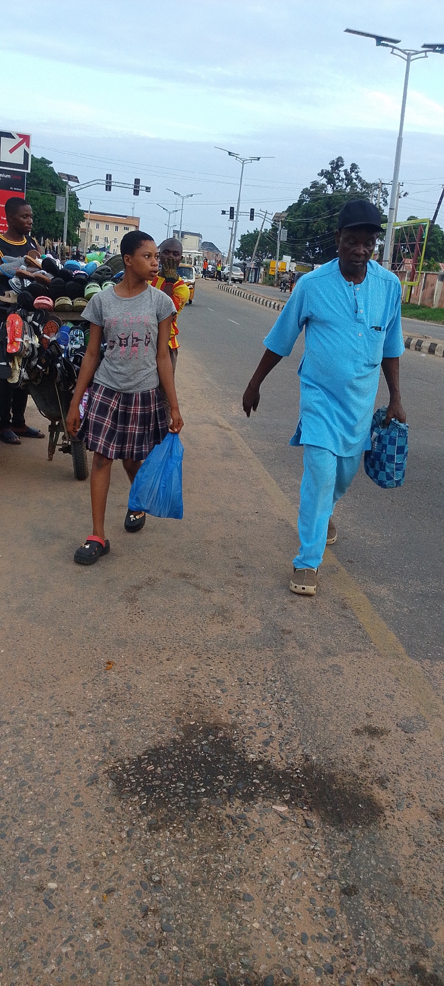The image captures a scene in Lokoja, Nigeria, on a paved road next to a sidewalk. A young woman in a grey shirt, plaid skirt, and black shoes walks along the road carrying a blue plastic bag. Beside her is a cart filled with an assortment of footwear, manned by a man. A man in a light blue two-piece outfit and a black cap walks in the opposite direction, carrying a checkered bag. The environment includes traffic lights, street lights, and other vehicles in the background, indicating an urban setting. The asphalt shows signs of wear and repair.