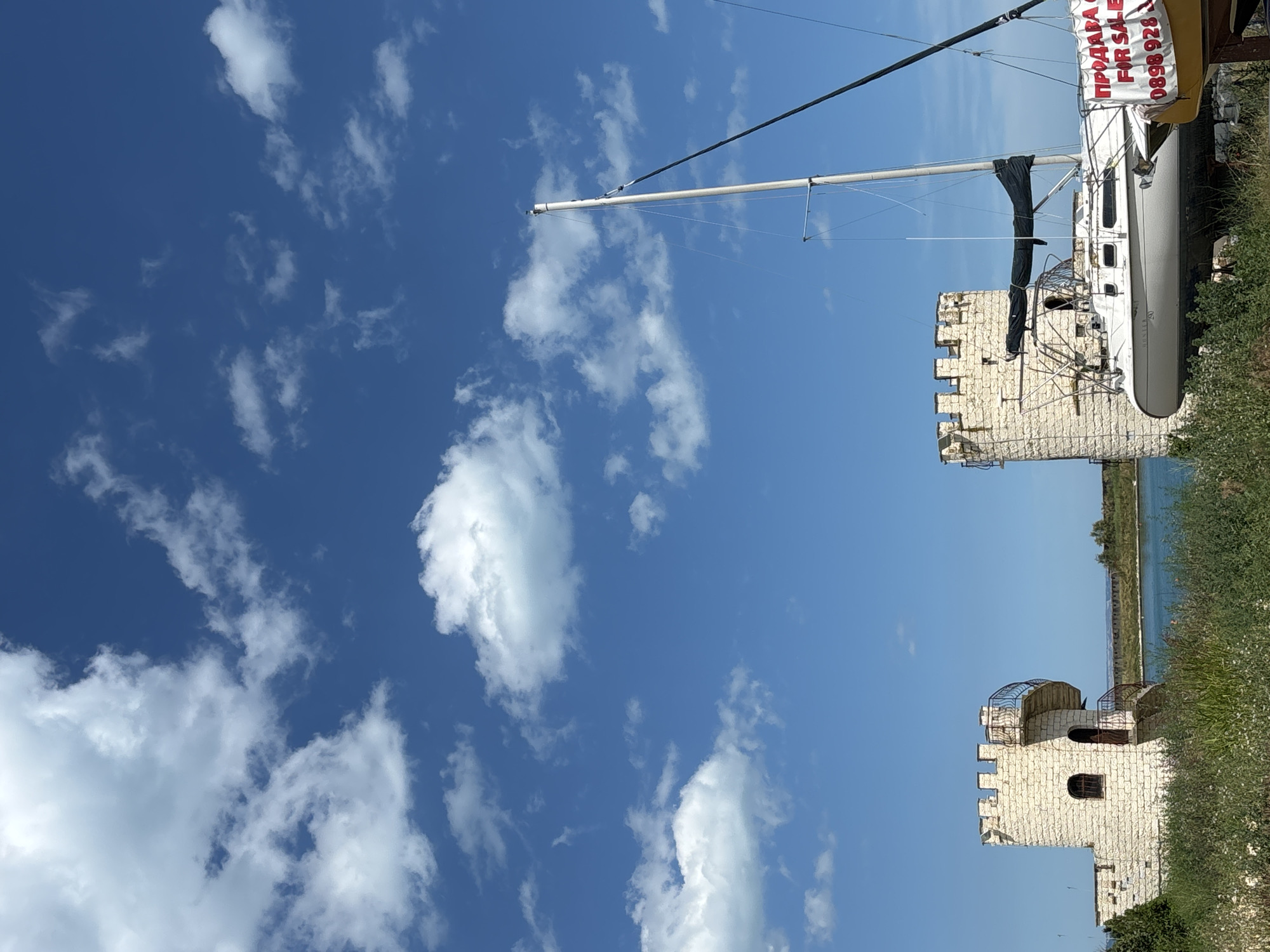 The media file shows a sailboat docked near a stone tower, with a clear blue sky and scattered white clouds in the background. The sailboat is white with a tall mast. A banner is draped over the side of the boat, displaying text in red. The stone tower has a crenellated top and arched windows. There is also a smaller, round tower with a metal railing visible. The scene appears to be a dry dock or a shore area. The lighting suggests it is daytime. The water is visible in the distance behind the towers and vegetation. The overall mood is serene and tranquil.
Visible text: "ПРОДАВА СЕ FOR SALE" and "0898 928 928".