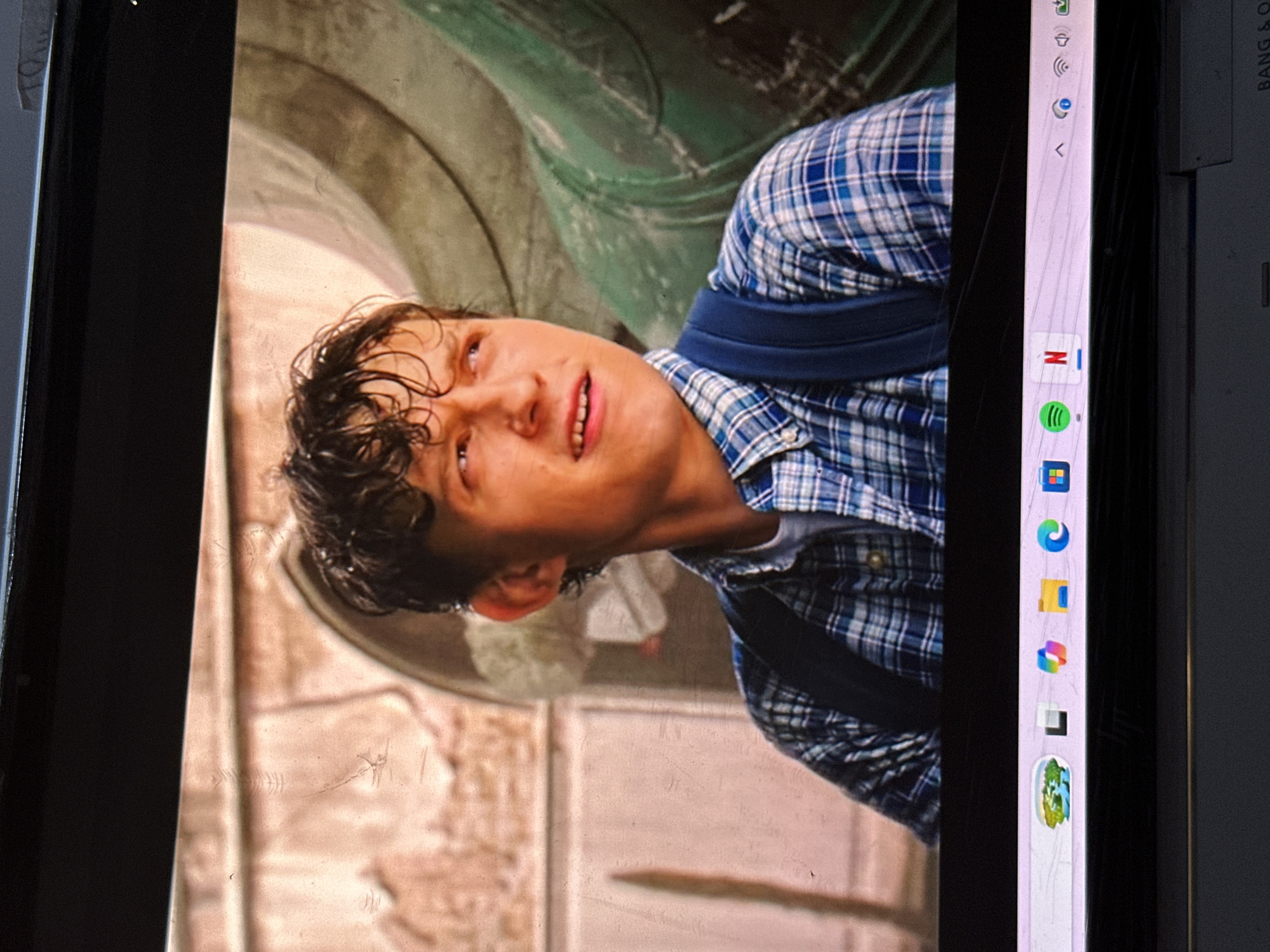 This media file is a photograph of a digital screen displaying a scene from a movie or TV show, captured in Bar Gada, Nigeria. The image is rotated 90 degrees clockwise.
The central subject on the screen is a young man, likely Tom Holland, with wet, curly brown hair. He is looking upwards and slightly to the side with his mouth slightly open, suggesting an expression of surprise, action, or mild distress. He is wearing a blue and white plaid shirt and a blue backpack. The background of the movie scene features old, weathered architectural elements, including large stone or concrete arches and walls in muted tones of green, grey, and pale brown.
To the right side of the screen's content, a vertical Windows taskbar is visible. It displays several icons, including those for Wi-Fi, volume, Netflix, Spotify, Microsoft Edge, File Explorer, a multi-colored square icon (possibly Teams or Paint 3D), and a distinct green and blue application icon at the bottom. The border of the screen, or bezel, is black, and part of a label, "BANG & O", is visible on the right side of the device, likely indicating the manufacturer (Bang & Olufsen). The screen surface itself shows some streaks and smudges, suggesting it has been used or handled.
The overall image captures the moment a person is watching content on a digital device, with elements of both the content and the device's interface and physical form visible.