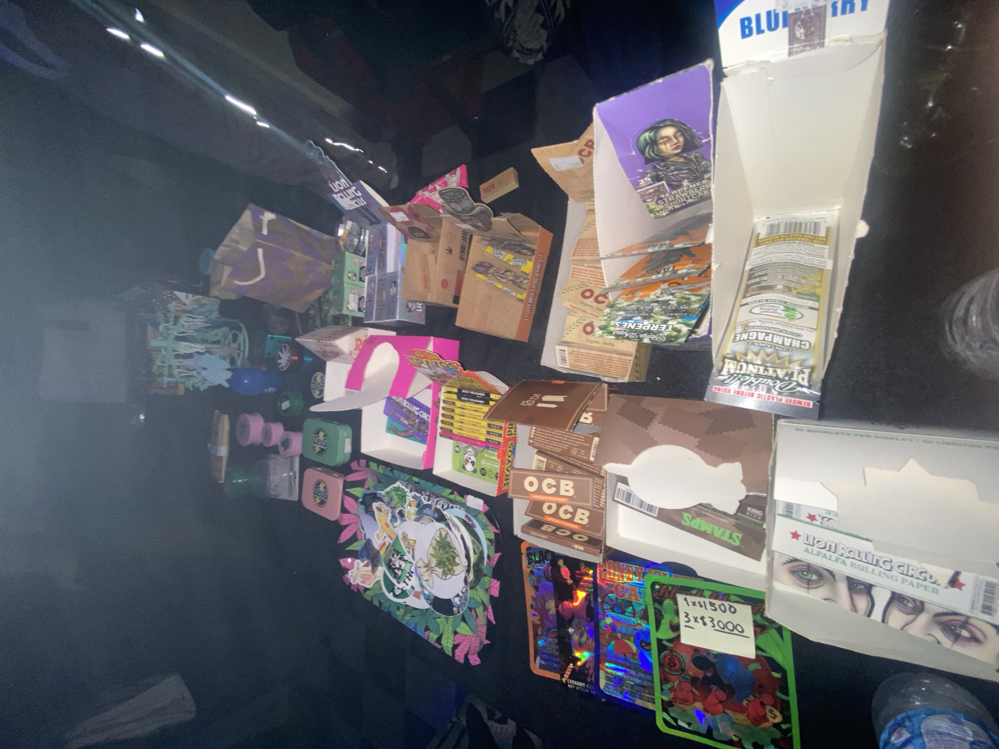 The image shows a collection of cannabis-related products and paraphernalia arranged on a black surface. The products include various brands of rolling papers, such as Lion Rolling Circus and OCB, as well as flavored blunt wraps (Champagne Platinum Double). There are also some small storage tins and decorative stickers with marijuana leaf designs. A handwritten sign indicates prices for an item, possibly related to the cannabis products. The scene appears to be set up for retail sale, or the organization of product for sale. It's impossible to ascertain its location within Buenos Aires, Argentina, without further contextual clues.