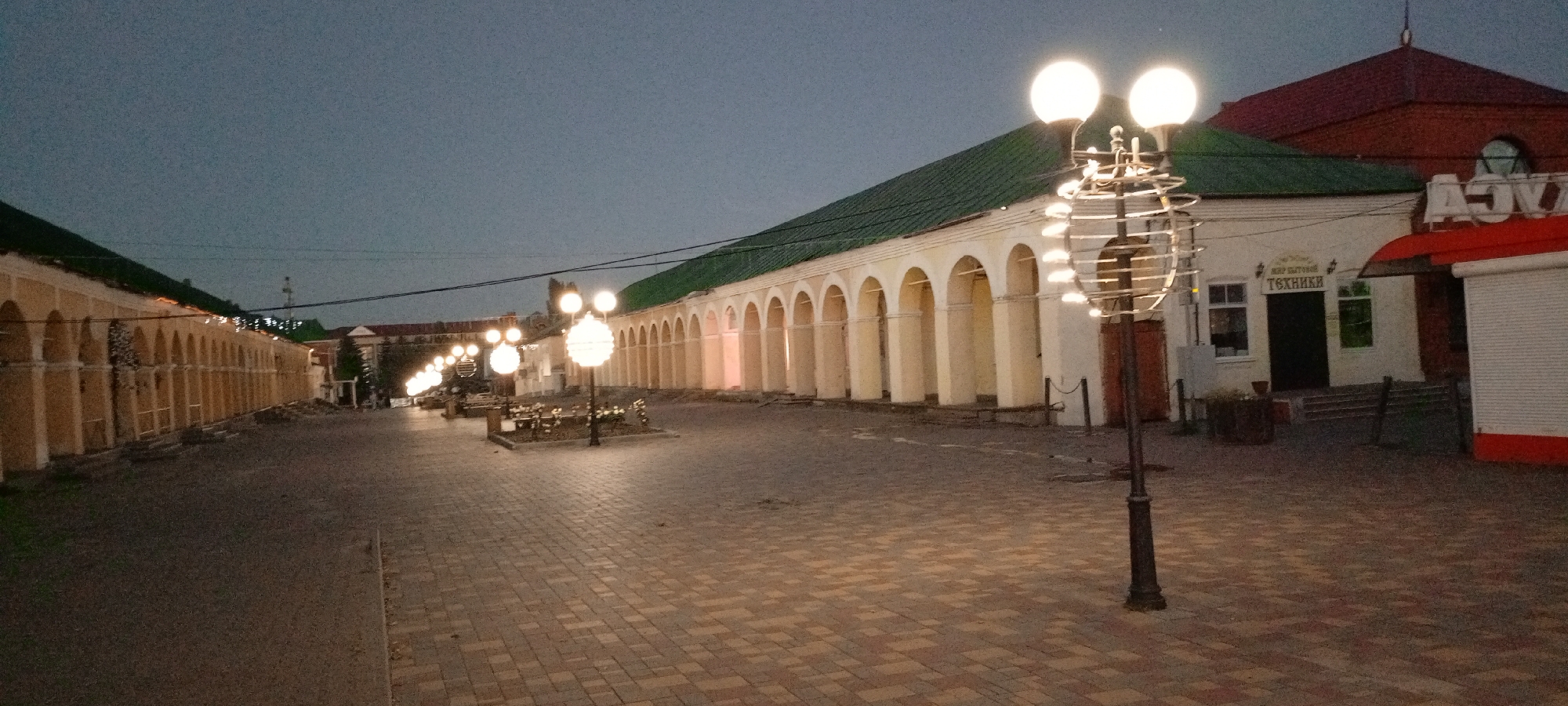 The image shows a street scene at dusk in Kirsanov, Russia. The street is paved with brickwork. On the left, there is a long building with yellow walls and a green roof, featuring a series of arches. On the right, there is another building with a red roof, a white facade, and a red awning. A sign with Cyrillic lettering is visible above an entrance. Several ornate street lamps with multiple illuminated globes line the street. In the foreground, a tall, dark lamppost with a decorative spiral element is prominent. There are no people visible in the image. The sky is overcast and dusky.