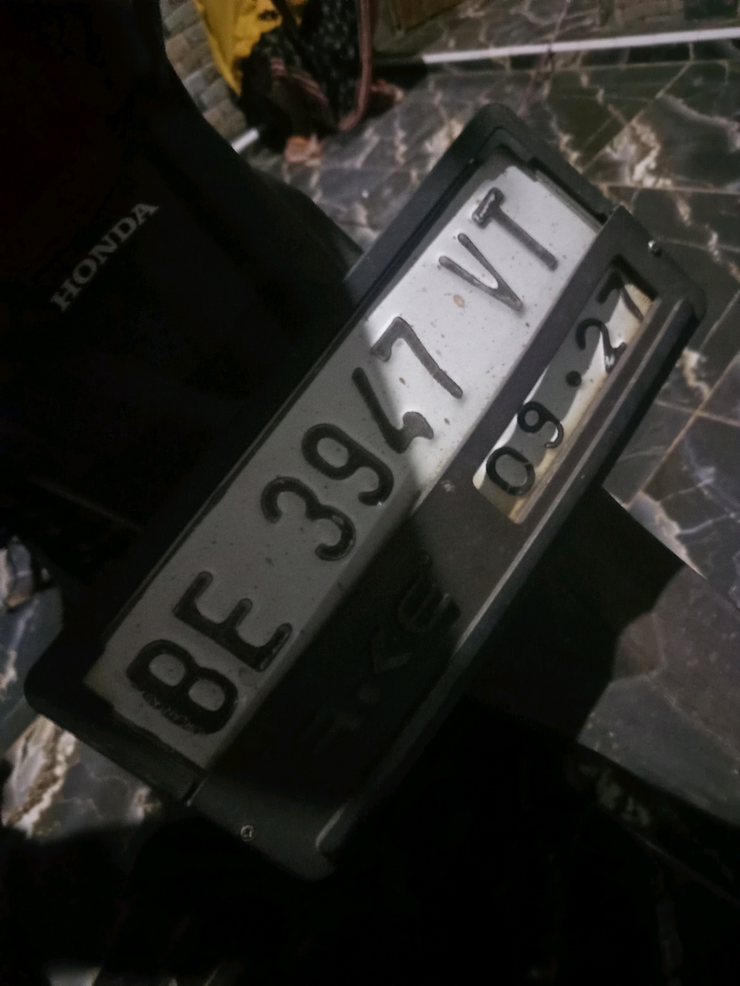 The image shows a close-up, angled view of a license plate on a motorcycle. The license plate reads "BE 3947 VT" and also displays "09.27". The license plate is mounted on a black frame, which is attached to the rear of a dark-colored motorcycle. The Honda logo is visible on the motorcycle's bodywork. The background is blurred but appears to be an outdoor setting with a patterned surface, possibly tiles, suggesting an urban environment. The lighting is dim, creating shadows and highlighting the reflective surface of the license plate. The image is taken at night or in a low-light condition. The location of Karang Sari, Indonesia, is not visually discernible from the image itself.