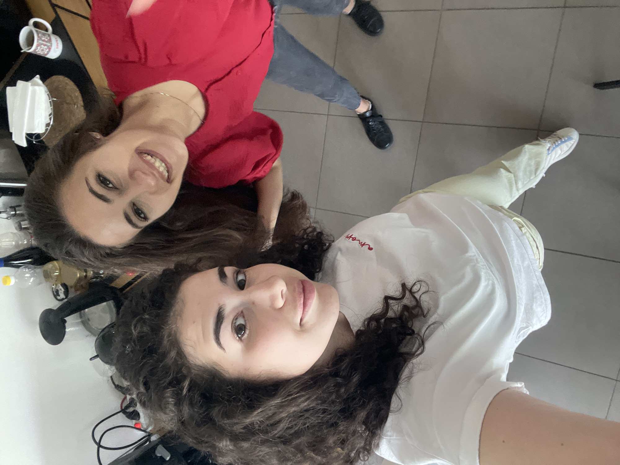 Two women are posing for a selfie. One woman is wearing a red shirt and grey pants and the other is wearing a white shirt and light yellow pants. The location is likely an indoor space with gray tile flooring, possibly a kitchen or similar area. There are items like a mug, napkins, and water bottles visible in the background, along with what appears to be kitchen equipment. The image does not reveal any information regarding the city of Sofia, Bulgaria.