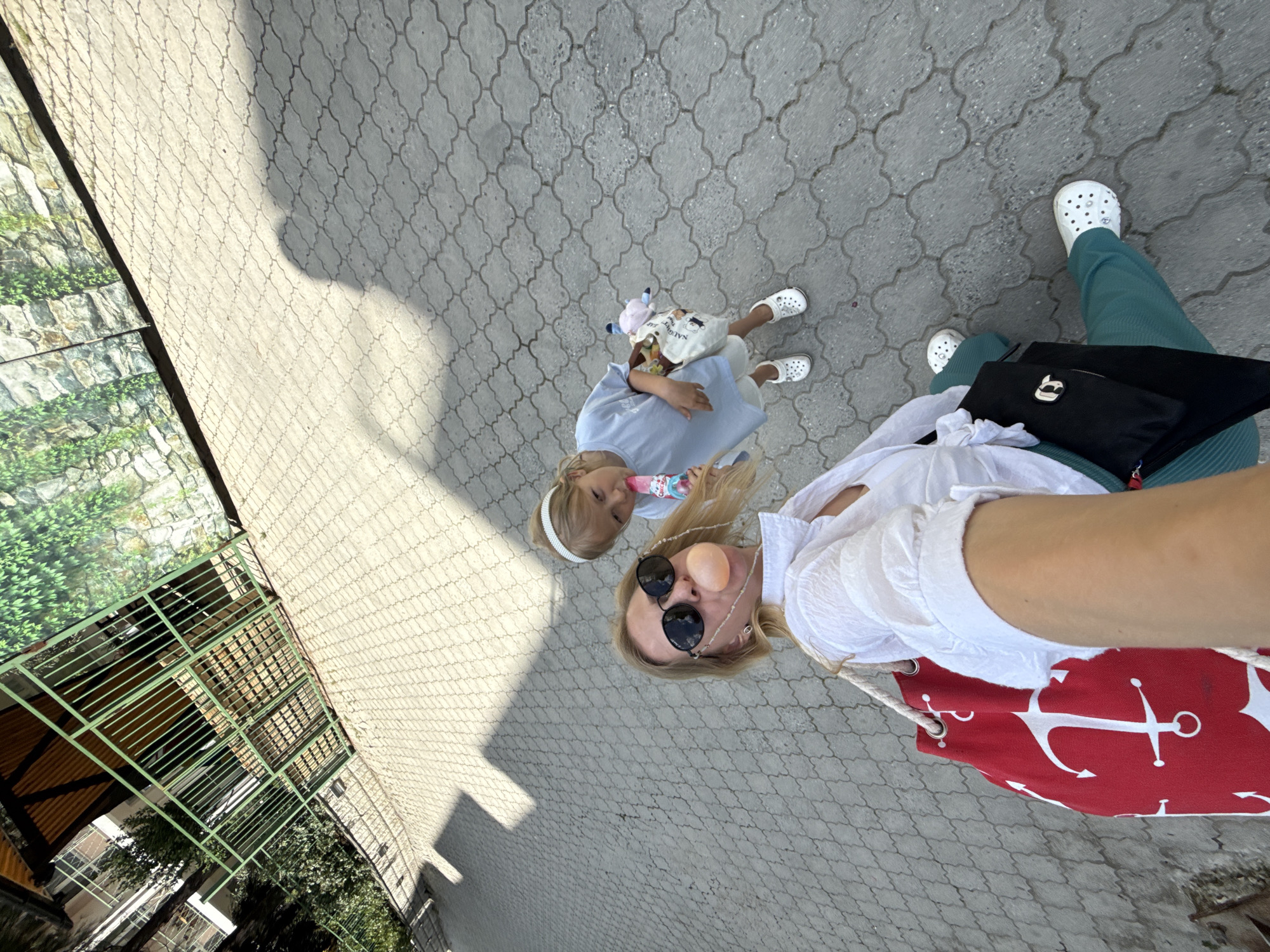 A low-angle, rotated shot shows a blonde-haired woman and a young girl walking on a gray paved surface in an urban environment. The woman, wearing sunglasses, a white blouse, and carrying a red and white anchor-patterned bag, has a gumball bubble on her face. The girl, in a light blue dress and headband, carries a plush toy. The background includes a green fence, the corner of a building, and a sunlit area.