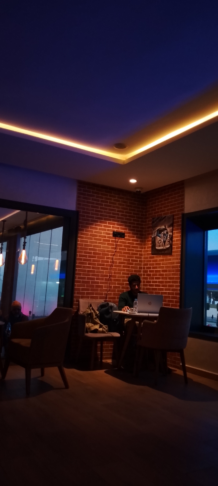 Inside a warmly lit space, a person sits at a small round table with a laptop. The backdrop is a brick wall adorned with a VW camper van poster. A window on the right shows a blue sky. To the left, another person sits in a chair near a window. The lighting is ambient, with concealed strip lighting on the ceiling. The room's ambiance is relaxed.