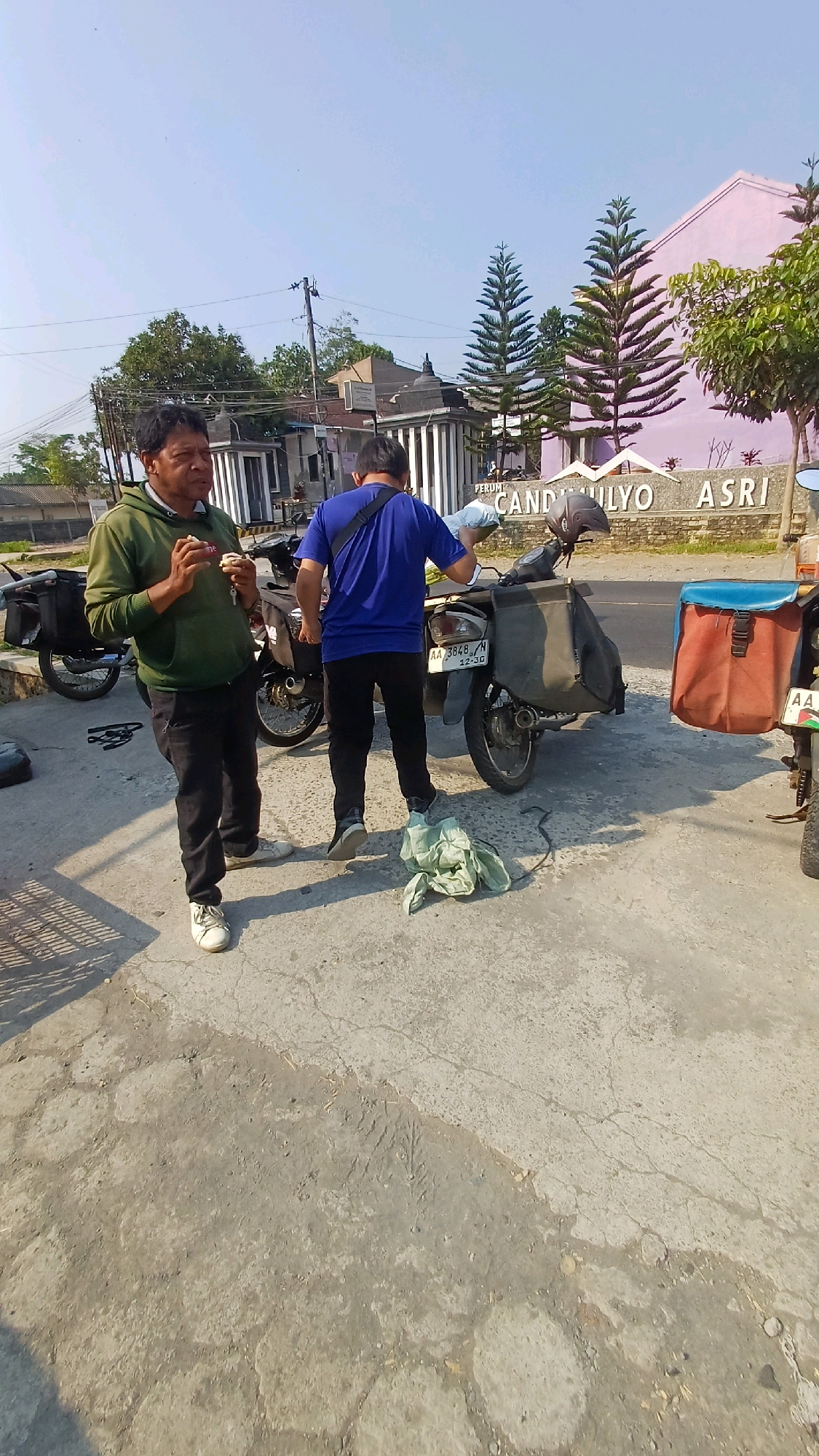 In the city of Kedu, Indonesia, under a clear blue sky, two men stand next to motorcycles on a paved area. The man on the left, wearing a green hooded jacket and black pants, is holding what appears to be food and looking down. The man on the right, dressed in a blue t-shirt and black pants, is facing away from the camera and interacting with a motorcycle. Two motorcycles are visible, one with black saddlebags and another with a dark green saddlebag and a brown and blue cargo box. A piece of light green cloth lies on the ground between the two men. In the background, a light pink building with decorative elements and text reading "PERUM CANDILYO ASRI" is visible, along with several tall, coniferous trees and power lines. The overall environment suggests an outdoor setting, possibly a residential area or a small business.