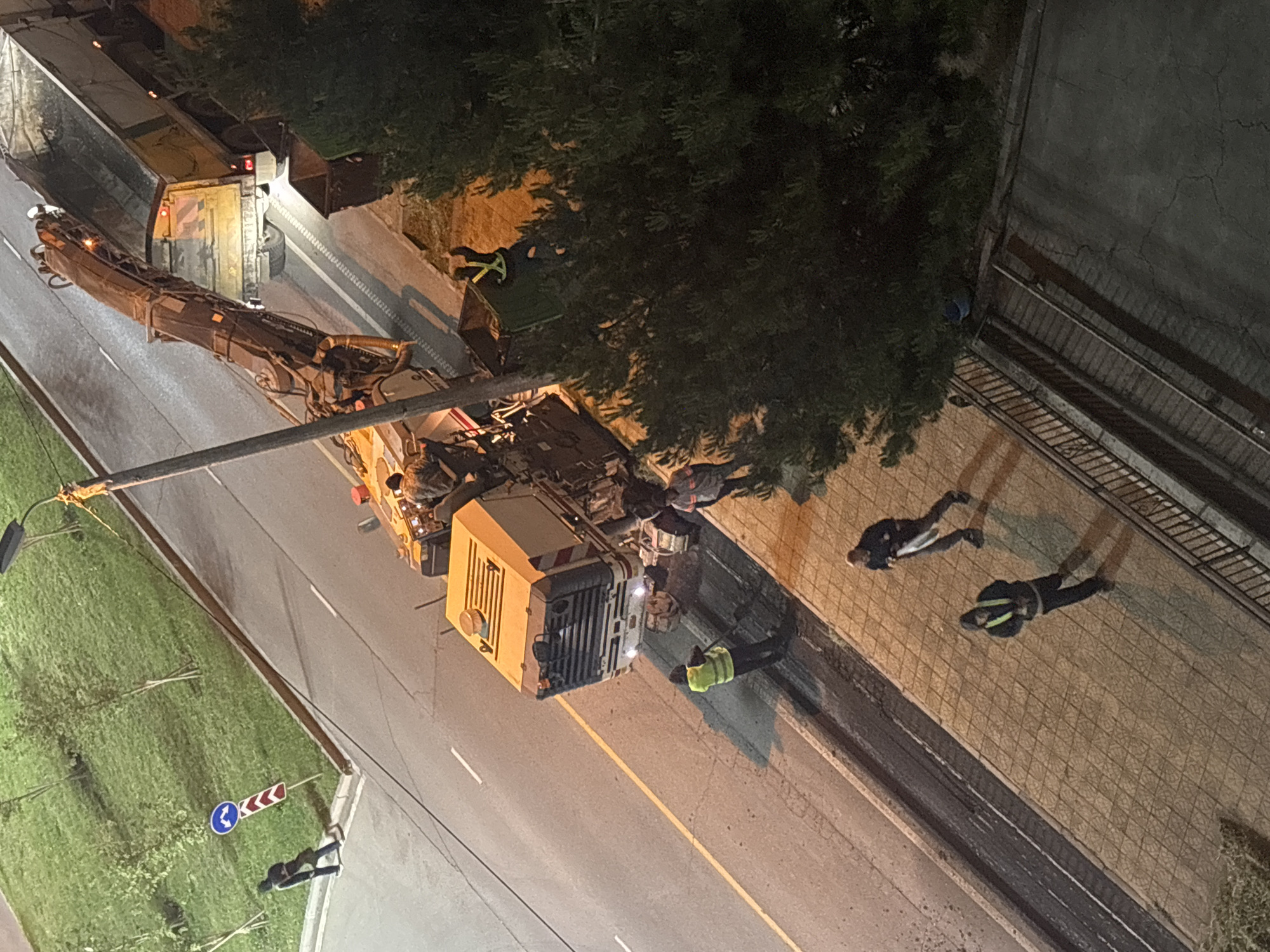 The image shows an aerial view of a road scene in Tbilisi, Georgia, at night. A large yellow truck with an extended crane arm is parked on the right side of the road, partially obstructing a lane. Several individuals are visible; one is near the front of the truck wearing a high-visibility vest, and two others are walking on the adjacent sidewalk. Vegetation, likely trees, are present on the right side of the road near the sidewalk. On the left side of the road, there is a green grassy area with a road sign visible.