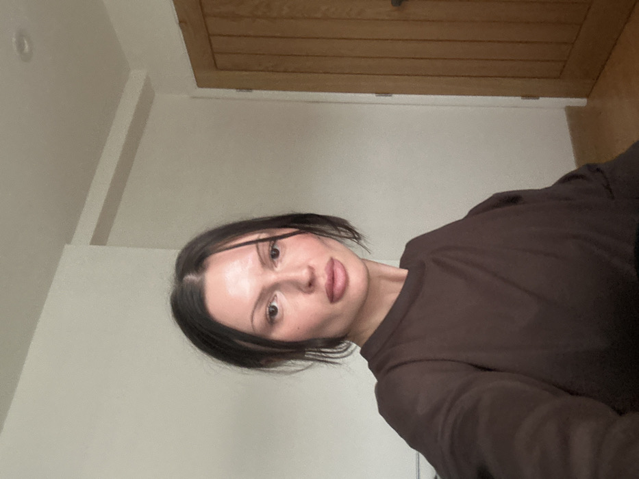 The image displays a female individual situated indoors, facing the camera with a neutral expression. She has dark, shoulder-length hair parted centrally and is wearing a dark brown long-sleeved garment. The background consists of a plain, light-colored wall. To the right, a light-toned wooden door featuring vertical panels and a dark handle is partially visible. A light-colored architectural trim runs along the ceiling line in the upper background. The scene is located within an unspecified indoor environment in Tunbridge Wells, United Kingdom.