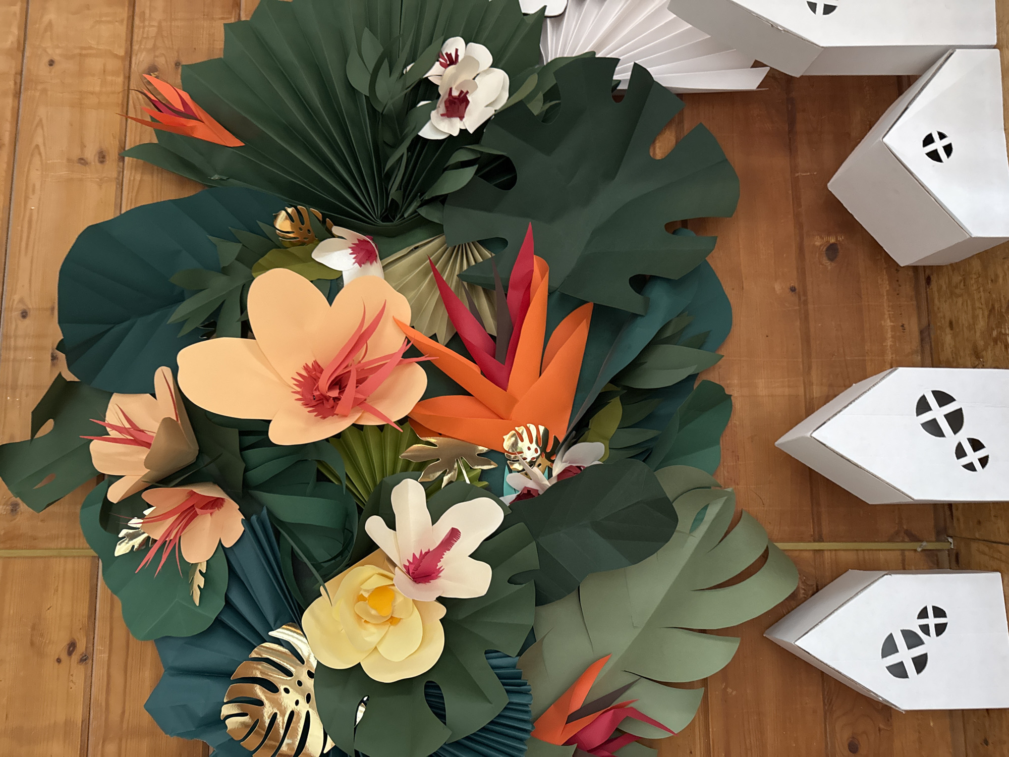 The image captures a vibrant, three-dimensional paper art installation displayed against a light brown wooden plank wall. The central focus is a dense, layered arrangement of paper-crafted tropical foliage and flowers, occupying the left and central parts of the frame.
The foliage consists primarily of large, dark green paper leaves, some intricately fan-folded to resemble palm fronds, and others cut out in the distinctive shape of Monstera deliciosa leaves. Interspersed among these green elements are various paper flowers: light orange/peach blossoms with detailed pink or red fringed centers, delicate white orchids accented with deep red, and a bright yellow multi-petaled flower. Additionally, striking orange and red paper "bird of paradise" flowers add a bold splash of color. Several metallic gold paper Monstera leaves are strategically placed, providing a shimmering accent.
To the right side of the image, vertically arranged against the wooden wall, are several white, house-shaped paper boxes. Each box features circular cutouts on its side, detailed with a black cross pattern, resembling stylized windows. The overall setting is an indoor decorative display. No people are visible, and there is no visible text within the frame. The scene is static, highlighting the intricate details and vibrant colors of the paper craftsmanship, creating a festive and exotic atmosphere.