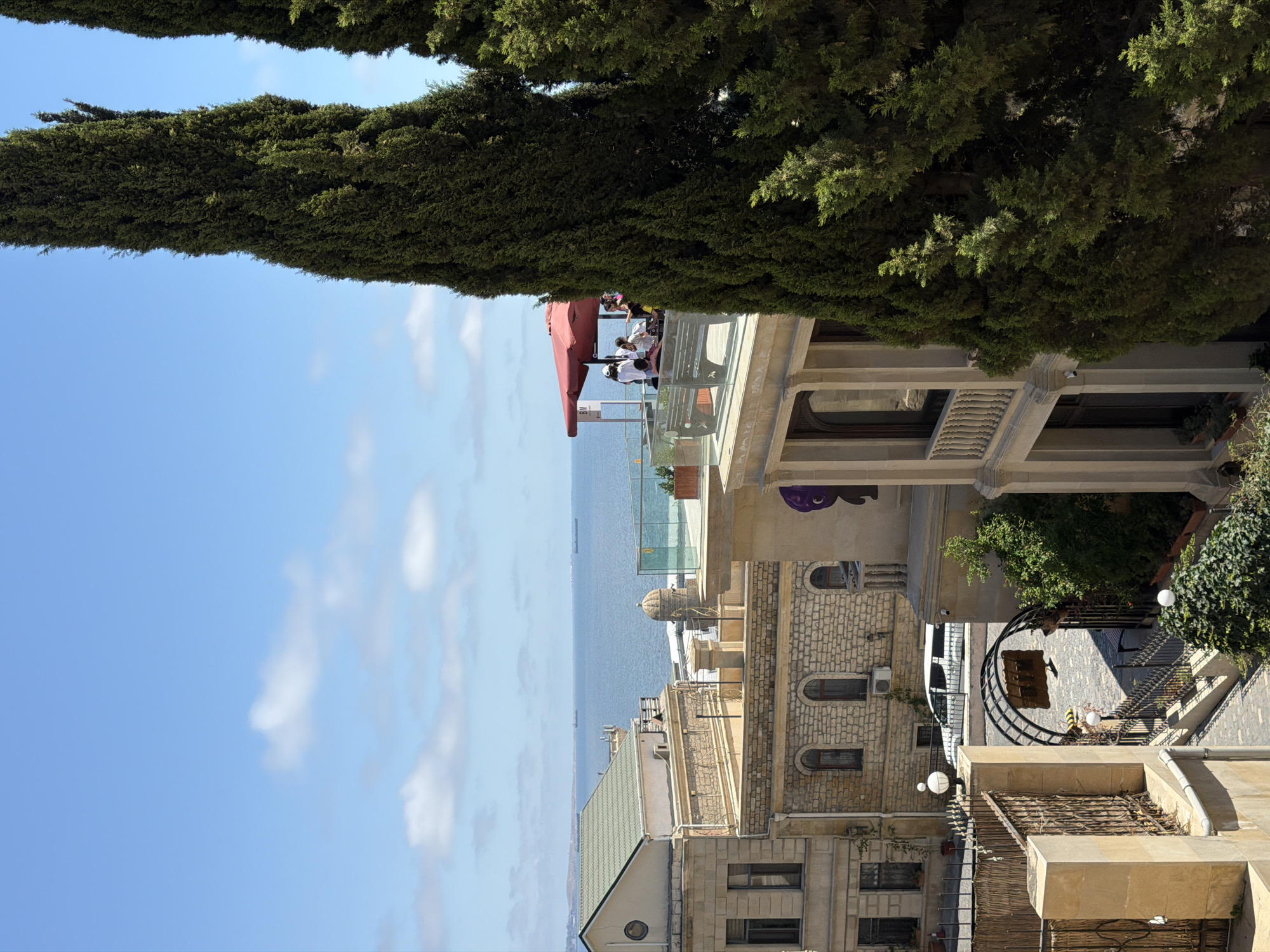 The image depicts a street scene in Sabail Raion, Azerbaijan, with a view of the Caspian Sea. In the foreground, two tall cypress trees frame the right side of the image. Behind them, a multi-story building with ornate architecture stands. This building features balconies with railings and large windows. To the left, another building constructed from stone is visible, with a green-tiled roof. Beyond these buildings, the Caspian Sea stretches out, with several ships visible in the distance under a bright blue sky dotted with white clouds. On a rooftop terrace of one of the buildings, a group of people are seated under a red umbrella, suggesting an outdoor seating area, possibly a cafe or restaurant. In the middle ground, a white vehicle is parked. The street area visible in the lower part of the image has cobblestone paving and is bordered by fences and decorative archways.
