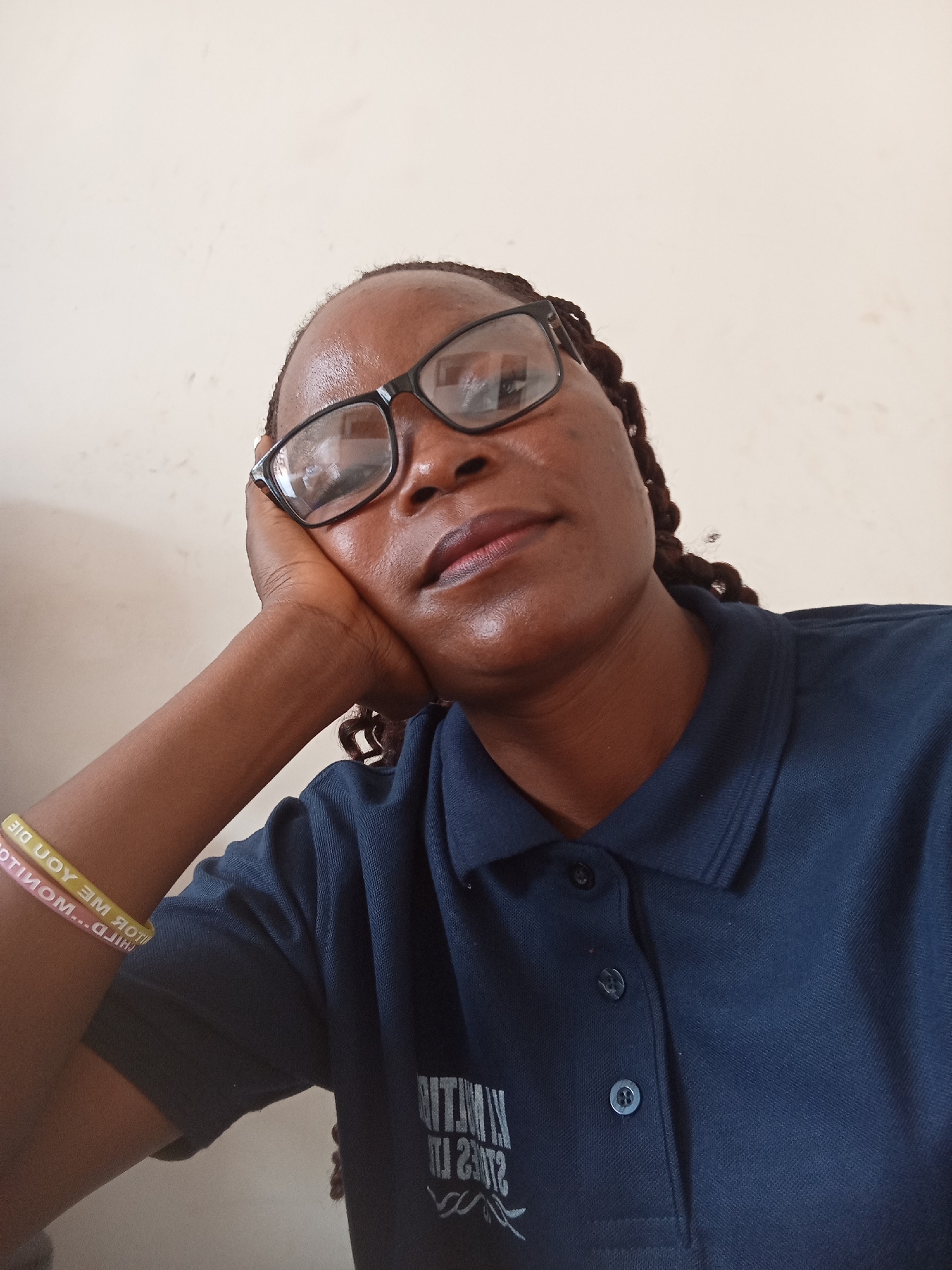 A close-up, low-angle, medium-shot photograph captures a Black woman resting her head on her hand, looking upwards. She is wearing glasses with black frames and a navy blue collared shirt with a two-button placket. The shirt has a white graphic emblem on the front. A braided section of her hair is visible near her right ear. On her left wrist, she wears two brightly colored rubber bracelets. The background consists of a plain, light-colored wall with minor imperfections. The lighting is soft, illuminating the subject's face and the immediate surroundings. The image appears to have been taken indoors, possibly in Jalingo, Nigeria, given the potential ethnic features of the subject.