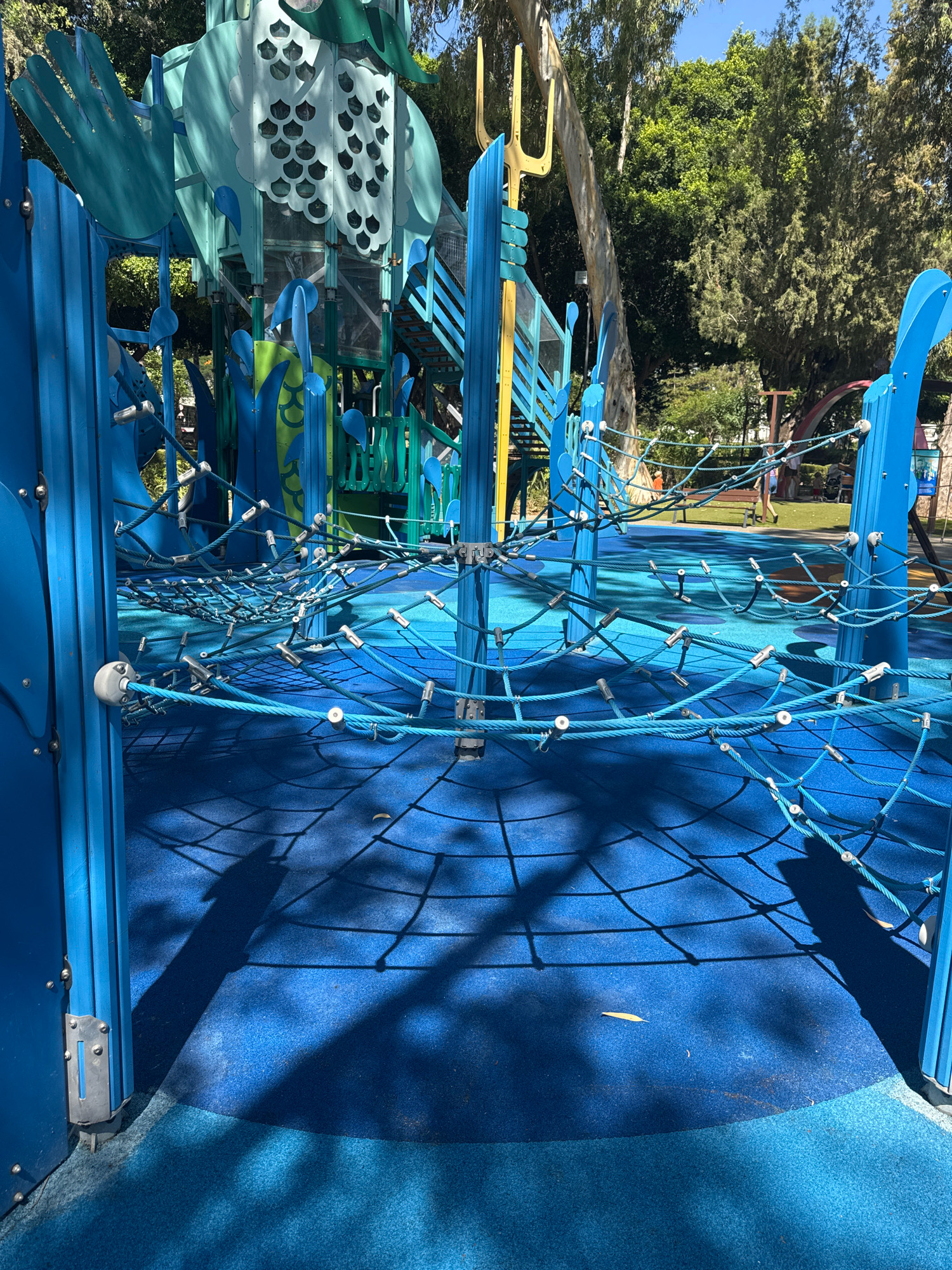 This image depicts a modern blue and green playground structure located in Limassol, Cyprus. The playground features a large, multi-level structure with slides, climbing nets, and colorful accents. In the foreground, a complex network of blue ropes forms a climbing net suspended over a blue rubber safety surface. Shadows from the ropes and playground structure are cast on the ground, indicating a sunny day. In the background, trees and other playground equipment are visible, along with a few people walking or standing in the distance. The overall scene suggests a well-maintained public recreational area.