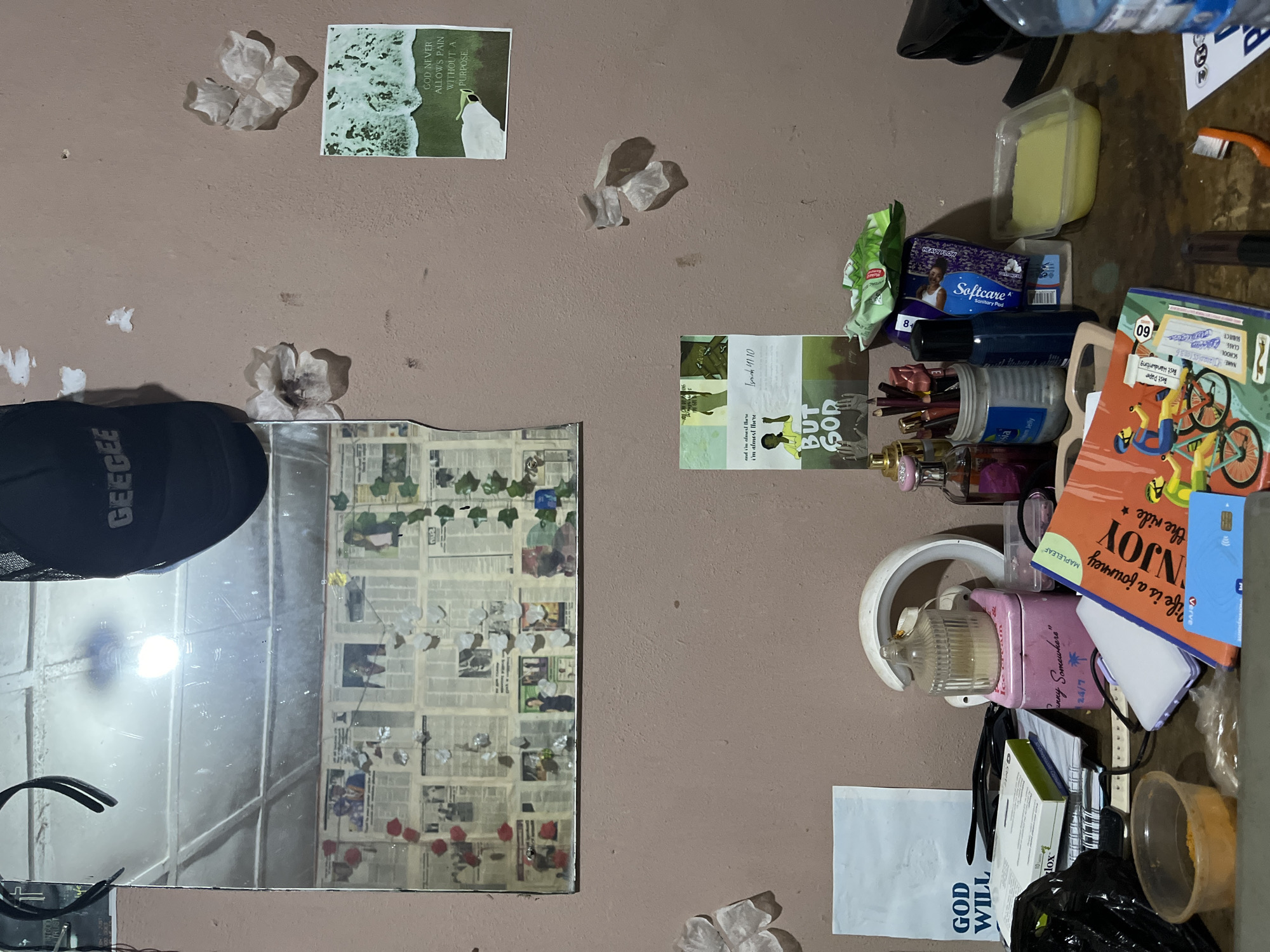 The image depicts a cluttered desk area within a room in Rafin Par, Nigeria. The wall behind the desk is painted a light brown and shows signs of wear, with several pieces of paper and what appear to be dried flower petals attached to it. A mirror above the desk is covered with newspaper clippings and more decorative petals.
On the desk itself, numerous objects are arranged haphazardly. These include a black baseball cap with the word "GEEGEE" visible, a black garbage bag, various containers, books, papers, pens, a bottle of perfume, and a bottle of water. A bright pink tin with the label "Funny Somewhere" and palm tree graphics is also prominent. A book with the title "ENJOY the ride" and illustrations of people cycling is open on the desk, along with a blue payment card. A transparent plastic container holds a yellow substance, possibly food.
The lighting suggests an indoor environment, likely a residential space, given the personal items and the general state of the room. The overall impression is one of a lived-in, personal workspace.