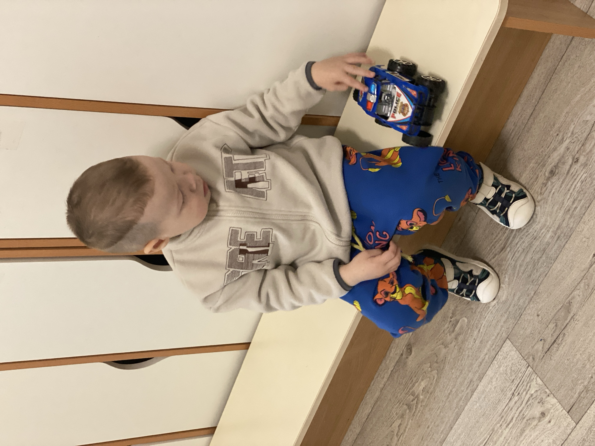 A young child, dressed in a light grey zip-up sweatshirt with the word "FREE" on it, blue Lion King print sweatpants, and white sneakers with dark blue accents, is sitting on a light beige surface next to a white dresser. He is playing with a blue and white toy police car with black wheels. The floor is light wood-grain vinyl. The location is unknown and there is no visible evidence to suggest the location is Sochi, Russia.