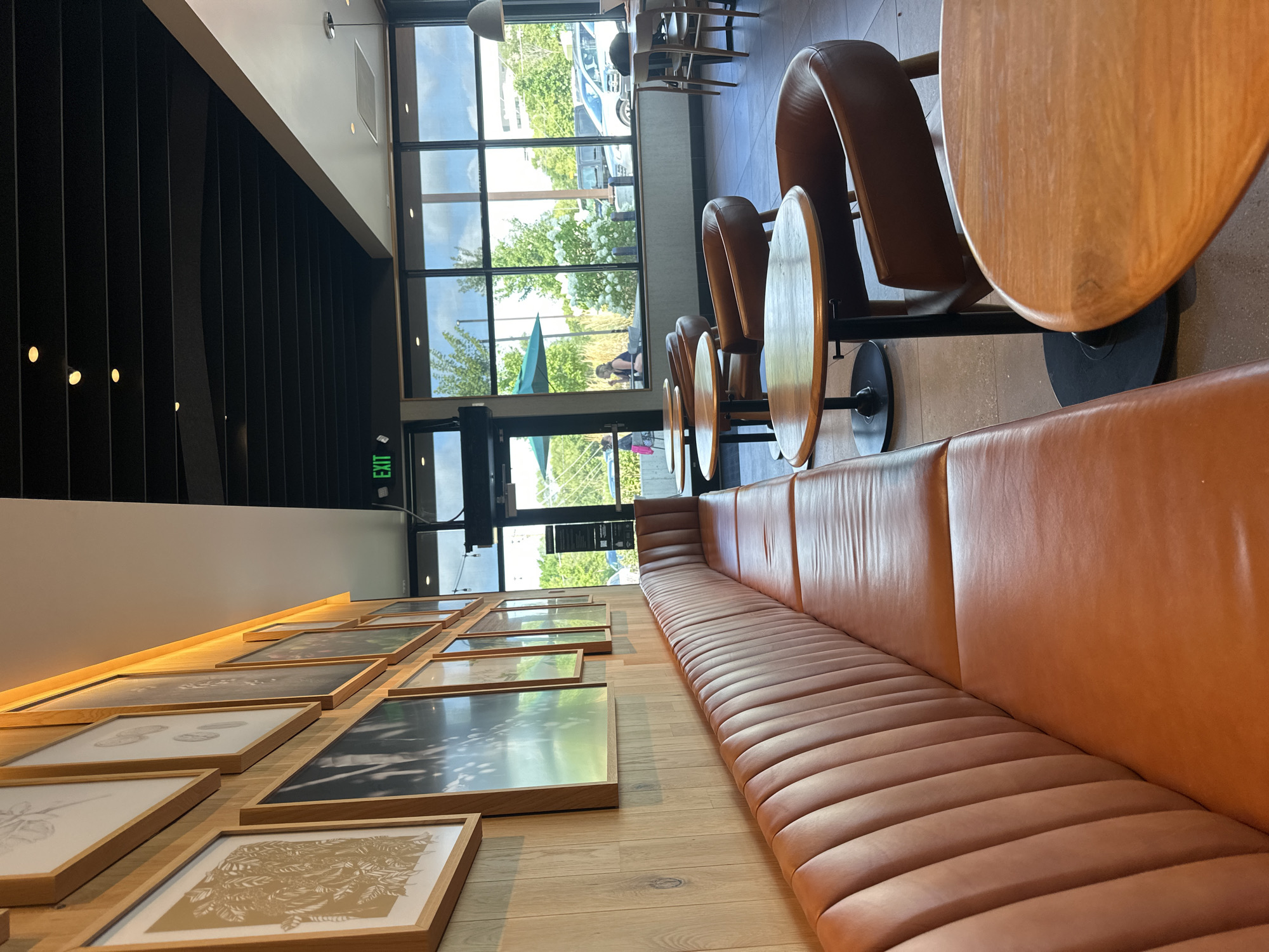 The image shows the interior of a Starbucks cafe located in McHenry, United States of America. The foreground features a long, brown leather booth with a ribbed design, typical of a cafe's seating. Several round wooden tables with black bases are scattered throughout the seating area. In the background, large windows offer a view of the outdoors, showing trees, cars, and a cloudy sky. The walls are decorated with framed artwork and wood paneling. An exit sign is visible above the windows. The overall ambiance is modern and comfortable, suggesting a typical Starbucks environment.