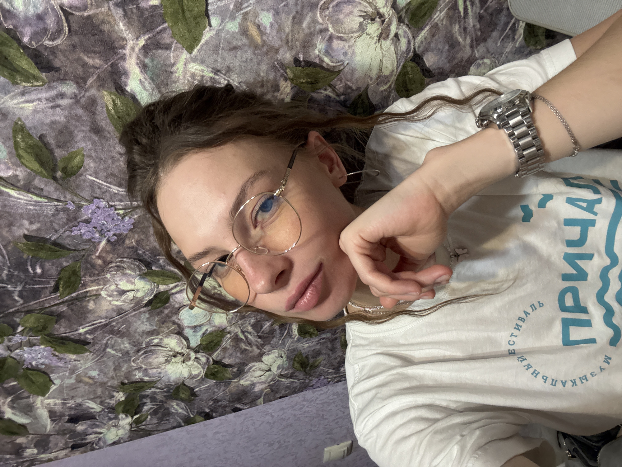 A woman with brown hair pulled back and wearing glasses is taking a selfie. She is wearing a white t-shirt with blue text on it and a silver watch on her left wrist. She is resting her chin on her hand. The background appears to be a floral wallpaper with shades of purple, green, and white. The lighting is dim, suggesting an indoor environment, possibly a residential setting. The image does not contain any identifiable landmarks or elements that would allow for specific localization within the city of Perm, Russia.