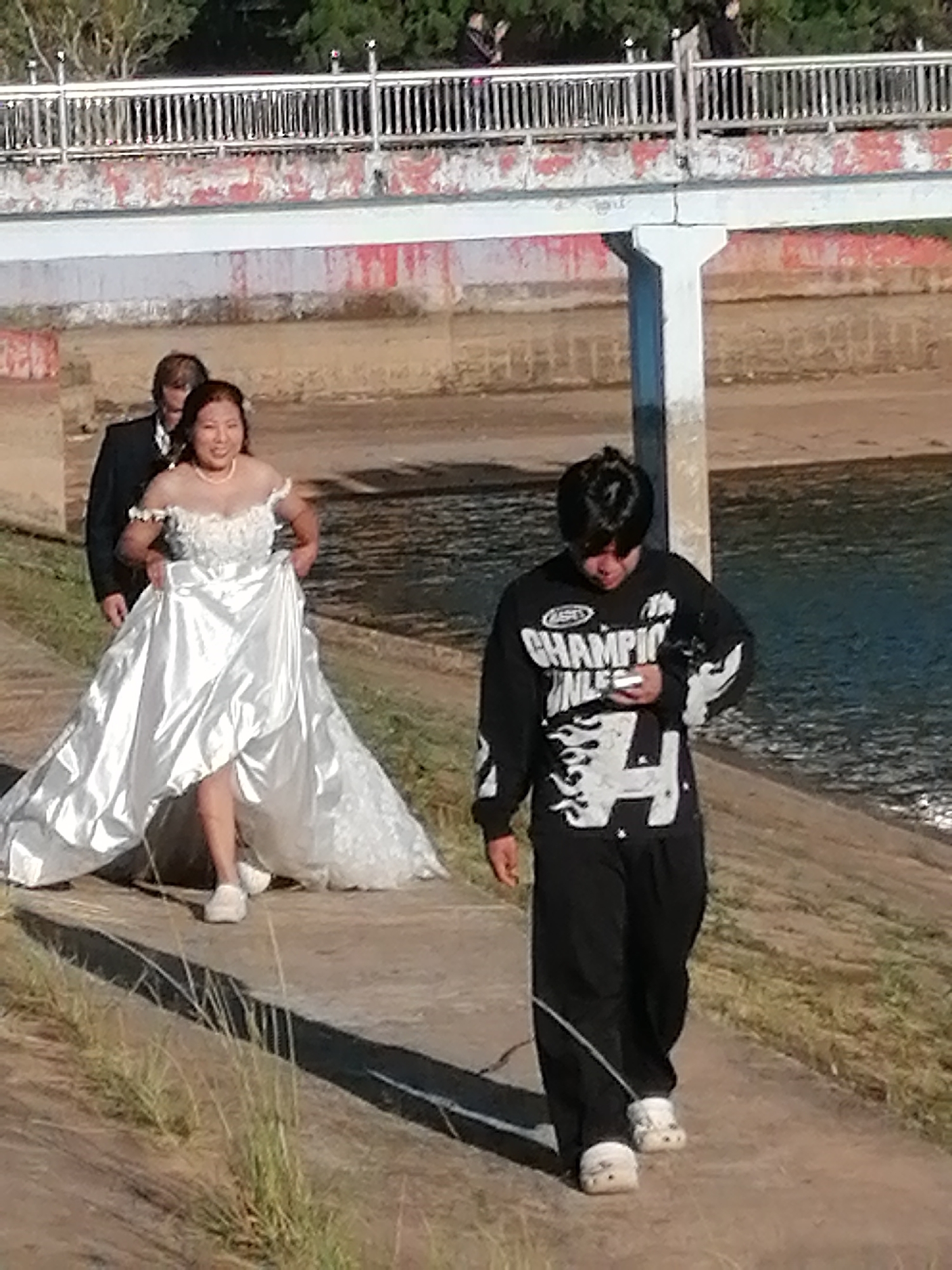The image captures a wedding party near a body of water, possibly the Xuan Huong Lake in Da Lat, Vietnam, judging by the landscape. In the foreground, a young person in a black long-sleeved shirt with a graphic design and black pants walks away from the camera, holding a white object, likely a phone, in their right hand. They are wearing white clogs. To the left, a woman in a white wedding dress with an off-the-shoulder neckline and a full skirt walks forward, holding the hem of her dress. She is wearing white shoes. Behind her and to the left, a man in a black suit and white shirt walks. In the background, a bridge with a metal railing and a concrete structure is visible, with some vegetation behind it. The environment suggests an outdoor event in a park or along a waterfront promenade.