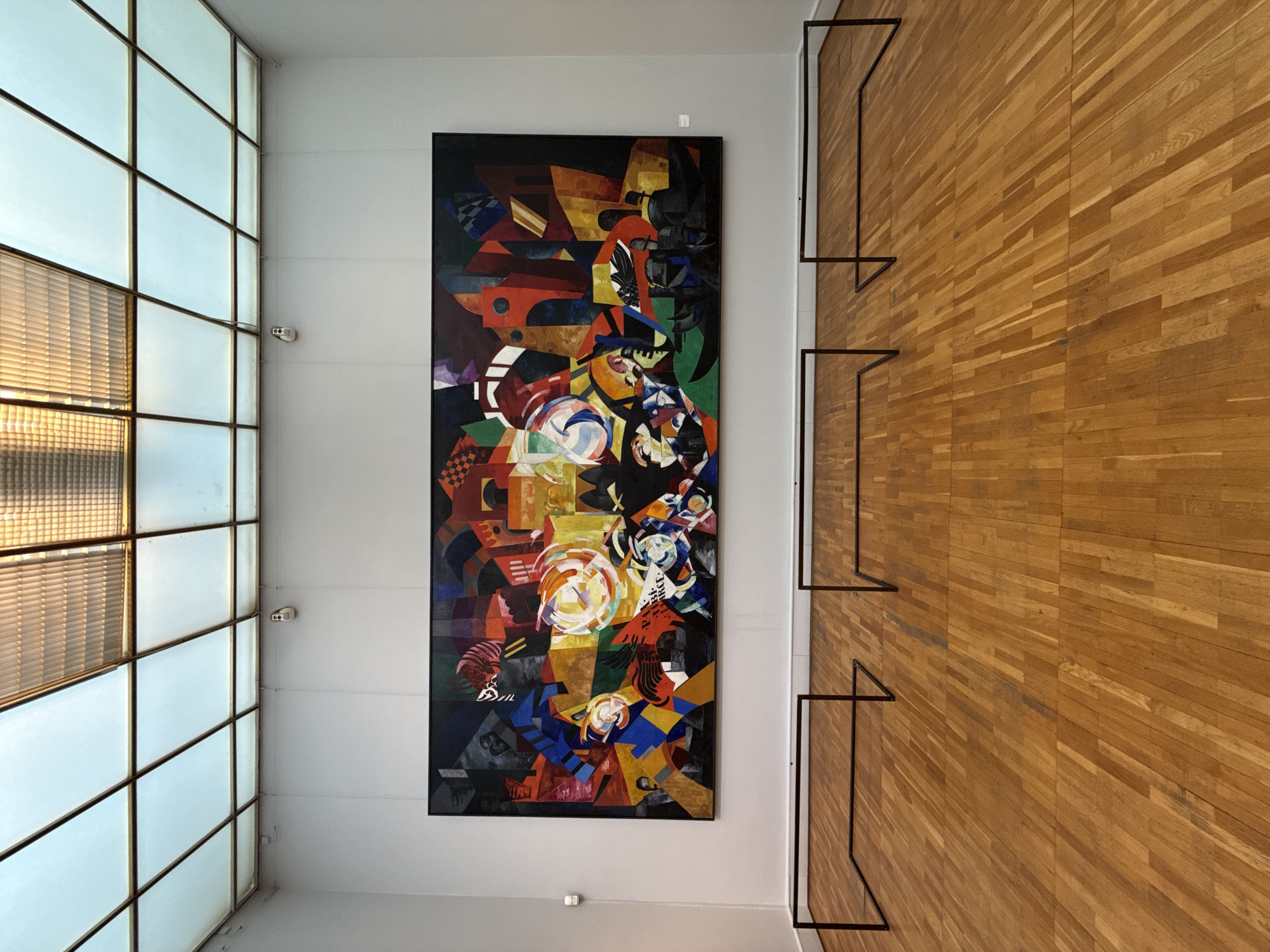 A photograph depicts an interior space with a tall window on the left and a wooden wall on the right. Centered between the window and the wall is a large, abstract painting with various geometric shapes and colors. Black metal frames are mounted on the wooden wall, possibly for display purposes. The window consists of a grid of glass panes with a partially visible blind behind it. The lighting appears to be a mix of natural light coming through the window and artificial light illuminating the scene.