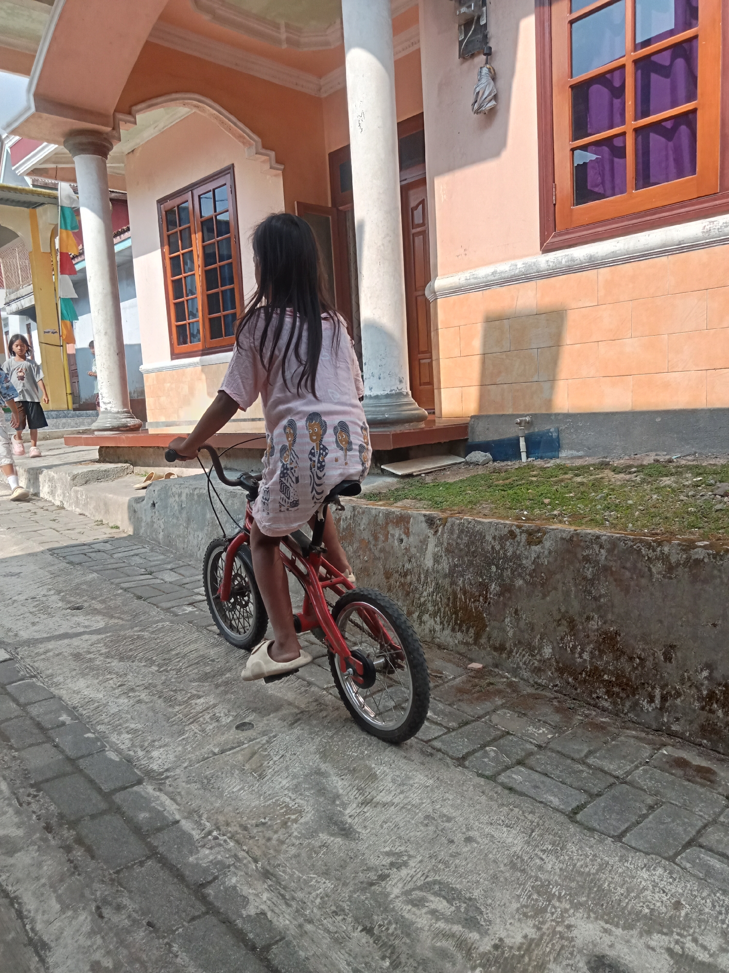 In Pancurwening, Indonesia, a young girl with long dark hair, wearing a light pink t-shirt featuring a pattern of figures, rides a small red bicycle. She is seen from behind, with her left hand on the handlebars and her right leg extended to pedal. She wears light-colored slip-on shoes. The bicycle has black tires with silver spokes.
She is on a paved pathway composed of gray rectangular bricks. To her left, a portion of a house is visible, characterized by a peach-colored exterior wall and a white pillar. A darker-colored door is also present. Further to the left, another young girl, wearing a gray t-shirt and dark shorts, stands facing forward.
Behind the girl on the bicycle is a raised concrete planter box with some green grass growing in it. Beyond this, the peach-colored wall of the house continues, featuring a dark brown window frame with multiple panes and purple curtains visible through the glass. A small section of a lighter-colored tiled wall is also present on the house's exterior. The environment appears to be an outdoor residential area.