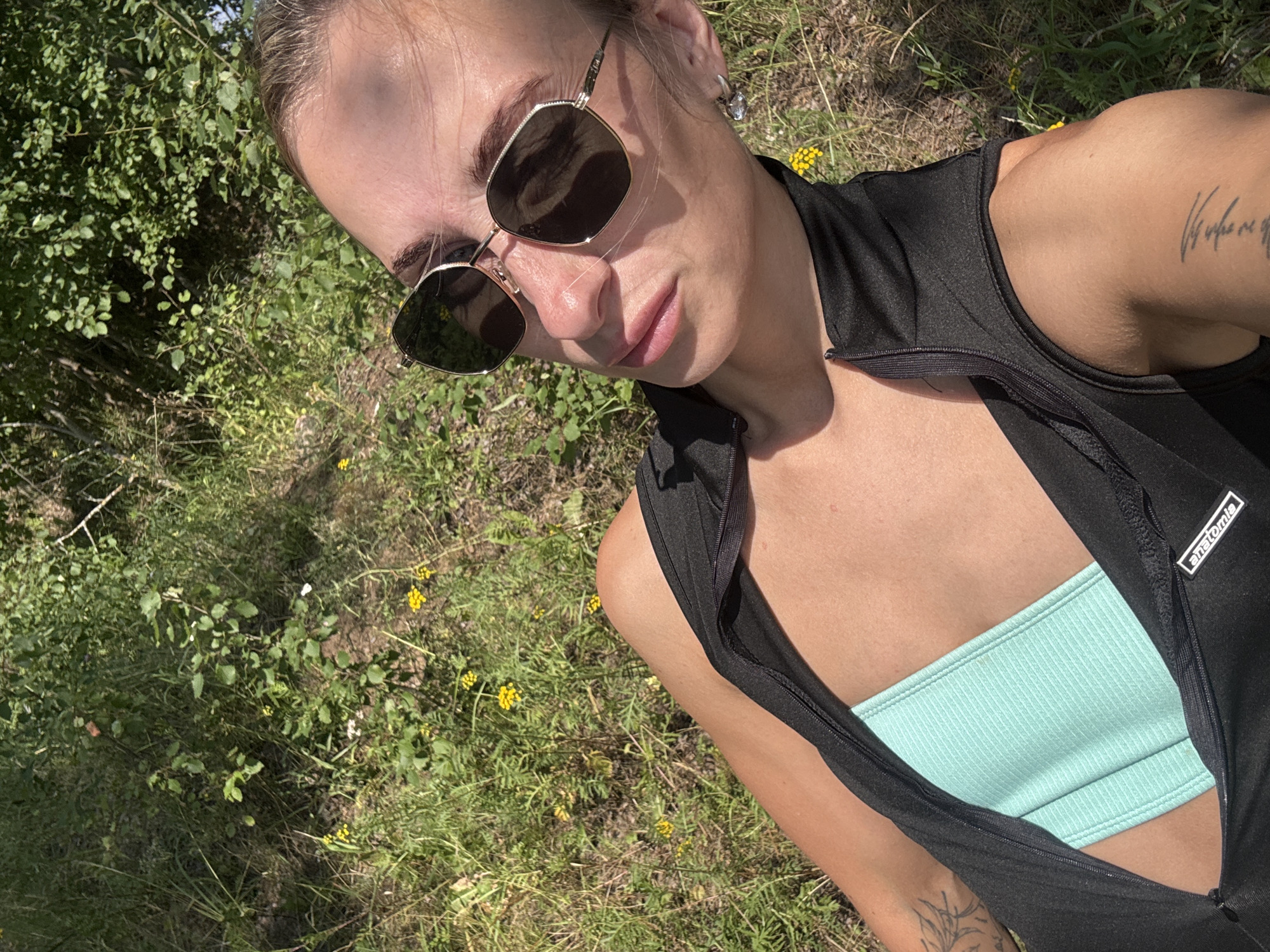 A young woman with brown hair, wearing sunglasses, a black zip-up vest, and a light blue top, lies in a grassy area with yellow wildflowers. She has tattoos on her arms. The scene appears to be outdoors in a natural, possibly rural setting. The image is a selfie taken from a high angle, looking down at the woman. The background consists of green foliage and dappled sunlight, suggesting it is daytime. No specific location in Noviy, Russia, can be identified from the image alone.