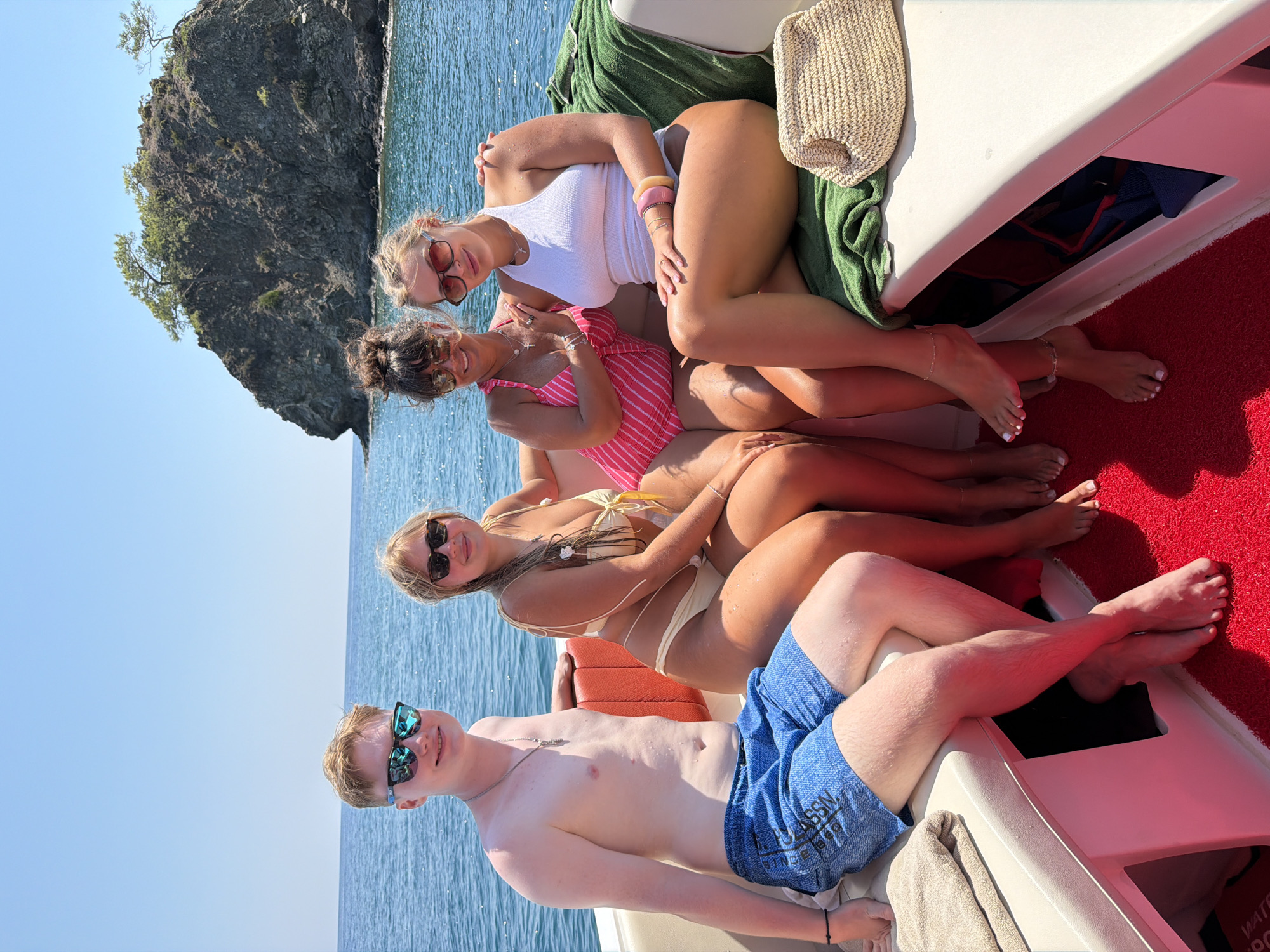 Four individuals, two males and two females, are seated on the deck of a boat under a clear blue sky. The boat has white seating with orange accents and a red carpeted floor. In the background, the turquoise sea is visible, with a rocky island topped with trees. The two females on the right are wearing swimsuits and sunglasses. One woman has on a pink and white striped one-piece swimsuit, and the other wears a white halter-neck tankini. The two individuals on the left are also dressed for a boat trip. The male on the far left is wearing blue patterned swim trunks and sunglasses, and a young female with blonde hair is seated next to him, wearing a light yellow bikini and sunglasses. The boat appears to be navigating the waters near Kemer, Turkey, given the distinctive rocky island formation visible in the Mediterranean Sea.