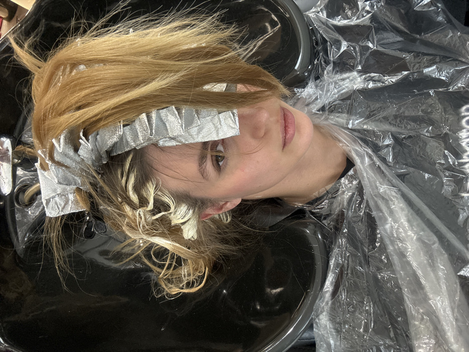 A female individual is depicted in an indoor hair salon, positioned with her head in a black hair wash basin. Her light brown and blonde hair is undergoing a chemical treatment, with multiple sections wrapped in silver foil, indicative of a highlighting or coloring process. Some hair sections appear bleached to a light blonde. Small black hair clips are interspersed throughout her hair. She wears a clear plastic protective cape over her torso, covering a dark garment on which the partial white text "Photon" is visible. The subject's face is partially obscured by the hair and foils, particularly her right eye and forehead. Her visible left eye is open, and she looks directly forward. The environment is consistent with a commercial hair styling establishment. A precise location within the city of Tbilisi, Georgia, is not identifiable from the visual information.