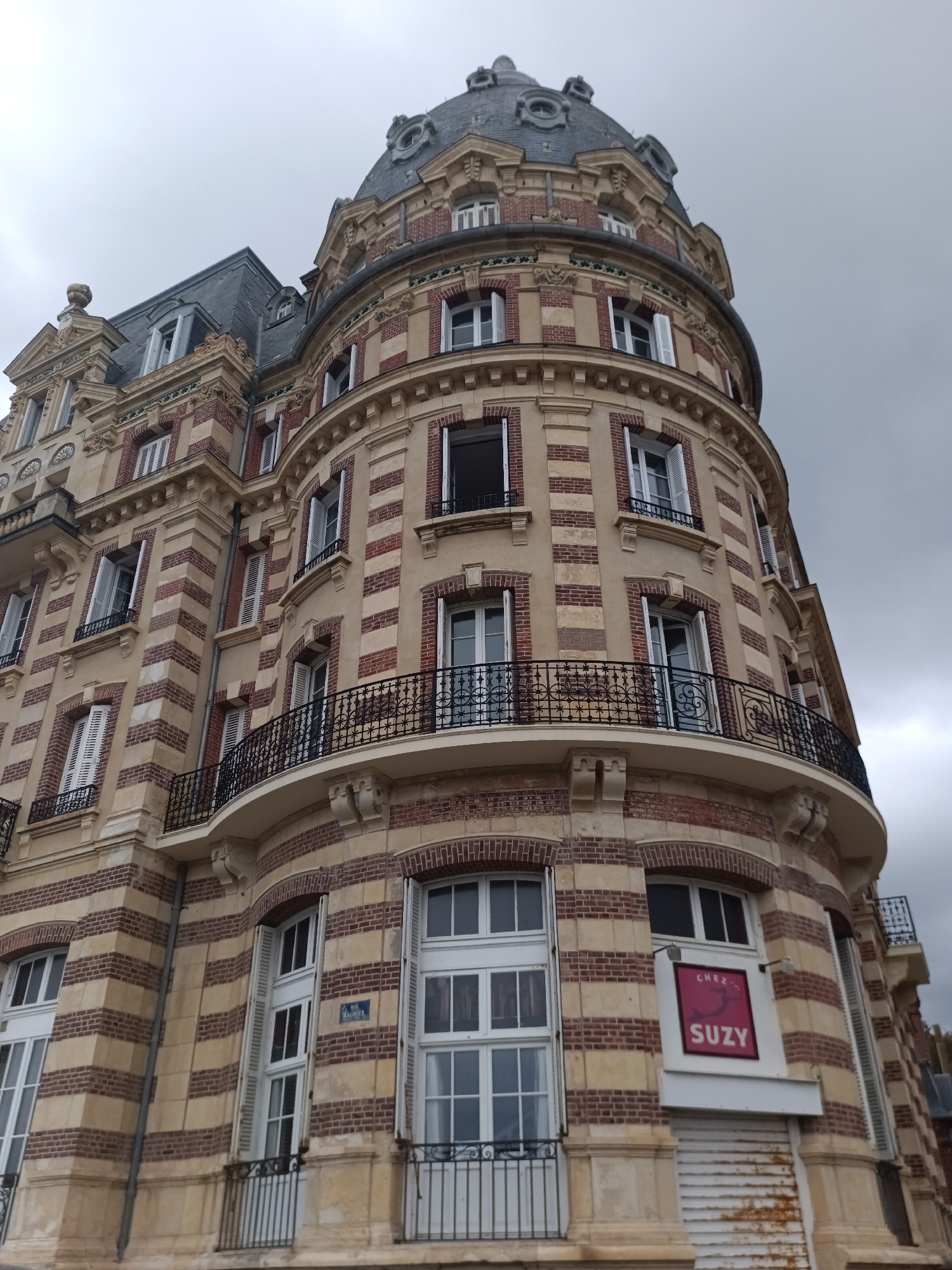 The image shows the exterior of a multi-story building in Houlgate, France, on an overcast day. The building features a distinctive rounded corner with balconies and red and beige striped brickwork. Windows with white shutters are visible on multiple floors. A sign with the text "CHEZ SUZY" is mounted on the lower section of the building, above a roll-up shutter. The architecture suggests a historic building, possibly a hotel or apartment complex. The sky is a uniform gray, indicating cloudy weather.