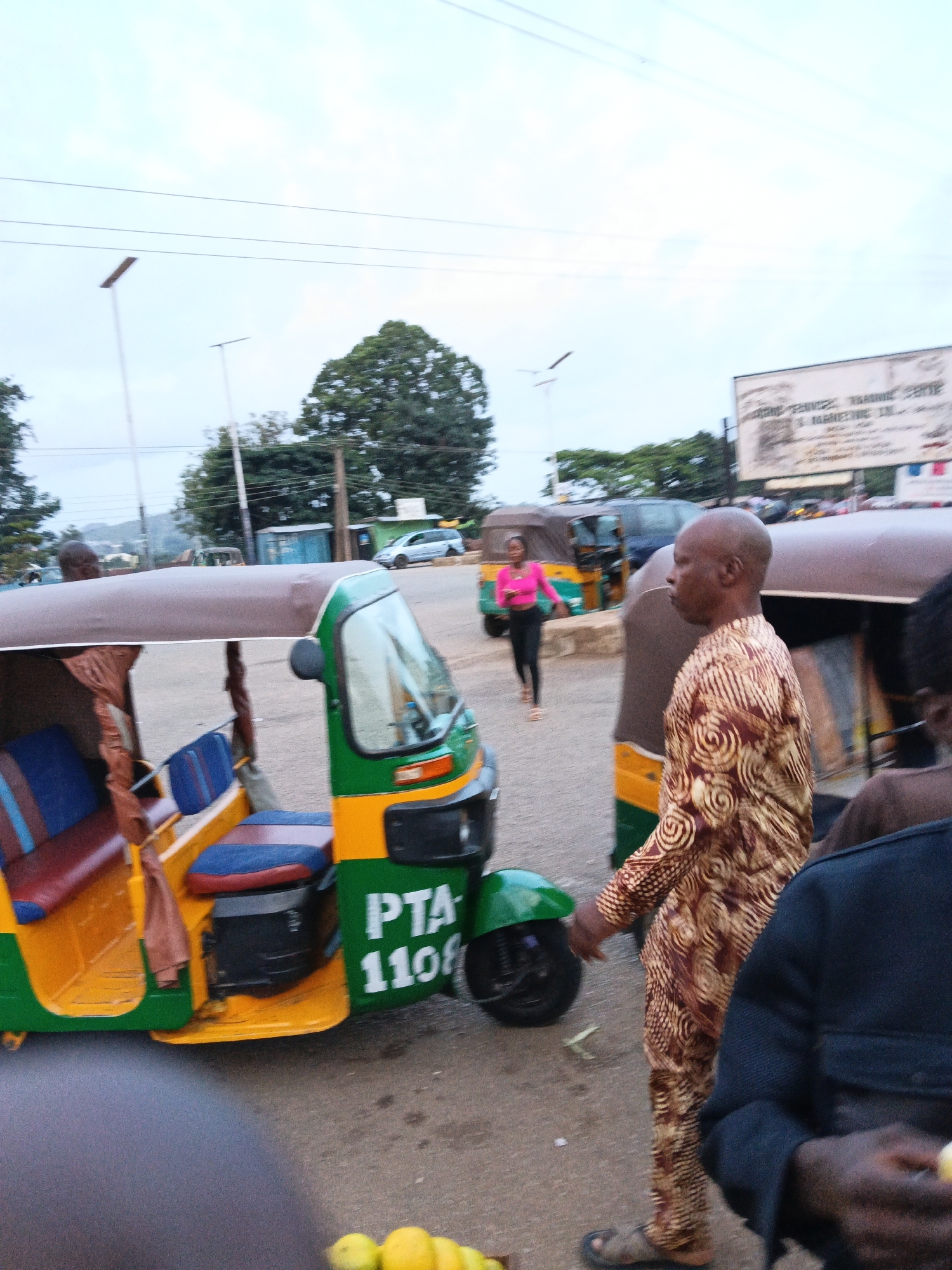 The image depicts an outdoor scene in what appears to be a city. In the foreground, a man wearing a patterned brown and gold outfit is walking. To his left, a green and yellow auto-rickshaw is parked. The auto-rickshaw has blue and red striped seating. The number "PTA 1108" is visible on the side of the auto-rickshaw. Further in the background, another auto-rickshaw and a woman in a bright pink top and black pants are visible. There are trees and buildings in the distance, and utility poles with wires are present. The sky is overcast. The overall scene suggests a street or roadside setting within a populated area.