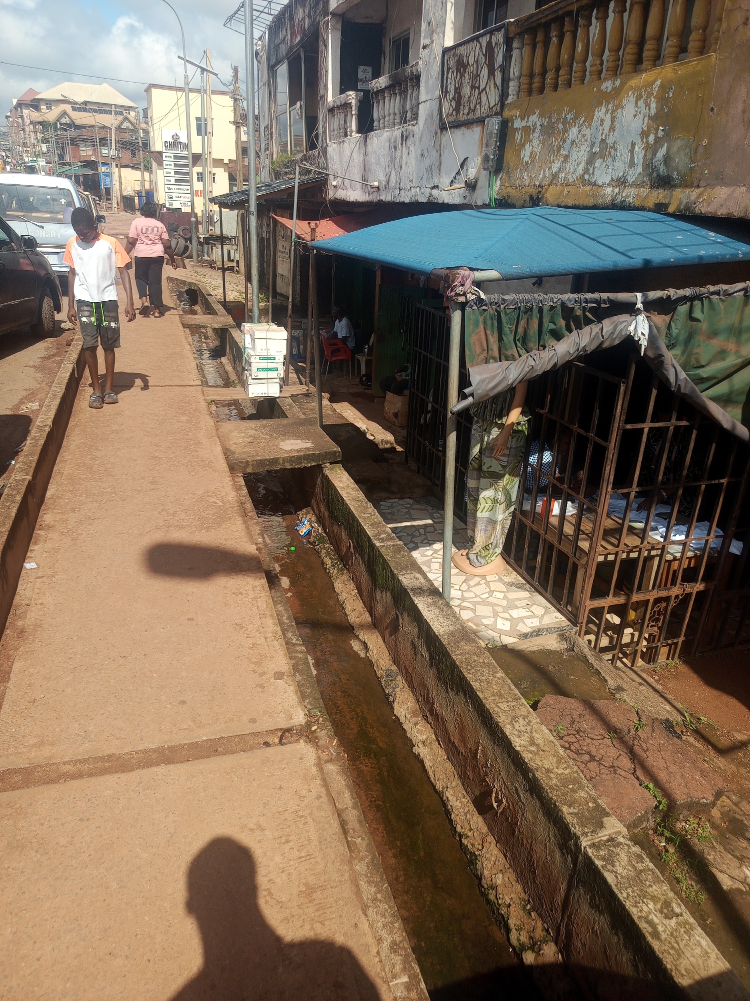 This is a medium shot of a street scene in Enugu, Nigeria, with a focus on a narrow walkway bordering a drainage channel. On the left, a young boy in an orange and white t-shirt and grey shorts walks away from the camera. Further down the walkway, a woman in a pink t-shirt and dark pants walks away from the camera. A white vehicle is parked on the left side of the street, partially visible.
The walkway itself is made of reddish-brown concrete. To the right of the walkway, a concrete drainage channel runs alongside, filled with murky water and some debris. Beyond the drainage channel, there is a structure with a blue tarp roof and metal bars, resembling a small shop or stall. Inside, a person in a patterned dress and another person are visible. In front of this structure, several stacks of white boxes are placed on the walkway.
The background shows a street lined with buildings of varying architectural styles and conditions. Some buildings have balconies with decorative railings. The overall impression is a busy, urban environment with signs of wear and tear. The sky is partly cloudy.