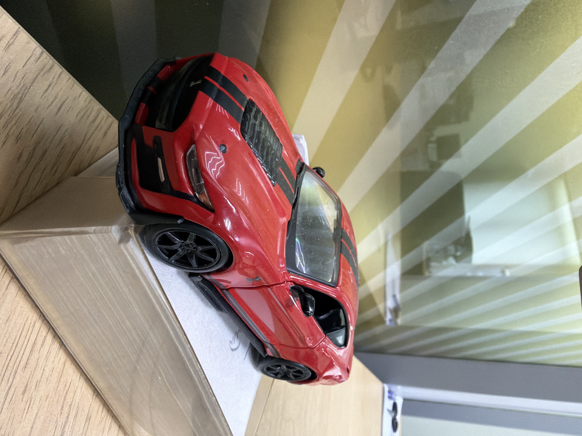 A red toy car with black racing stripes sits on a cardboard box. The car is on the corner of the box with part of the car overhanging. The background is a wall with yellow and white horizontal stripes. The location is not provided in the prompt and therefore cannot be determined.