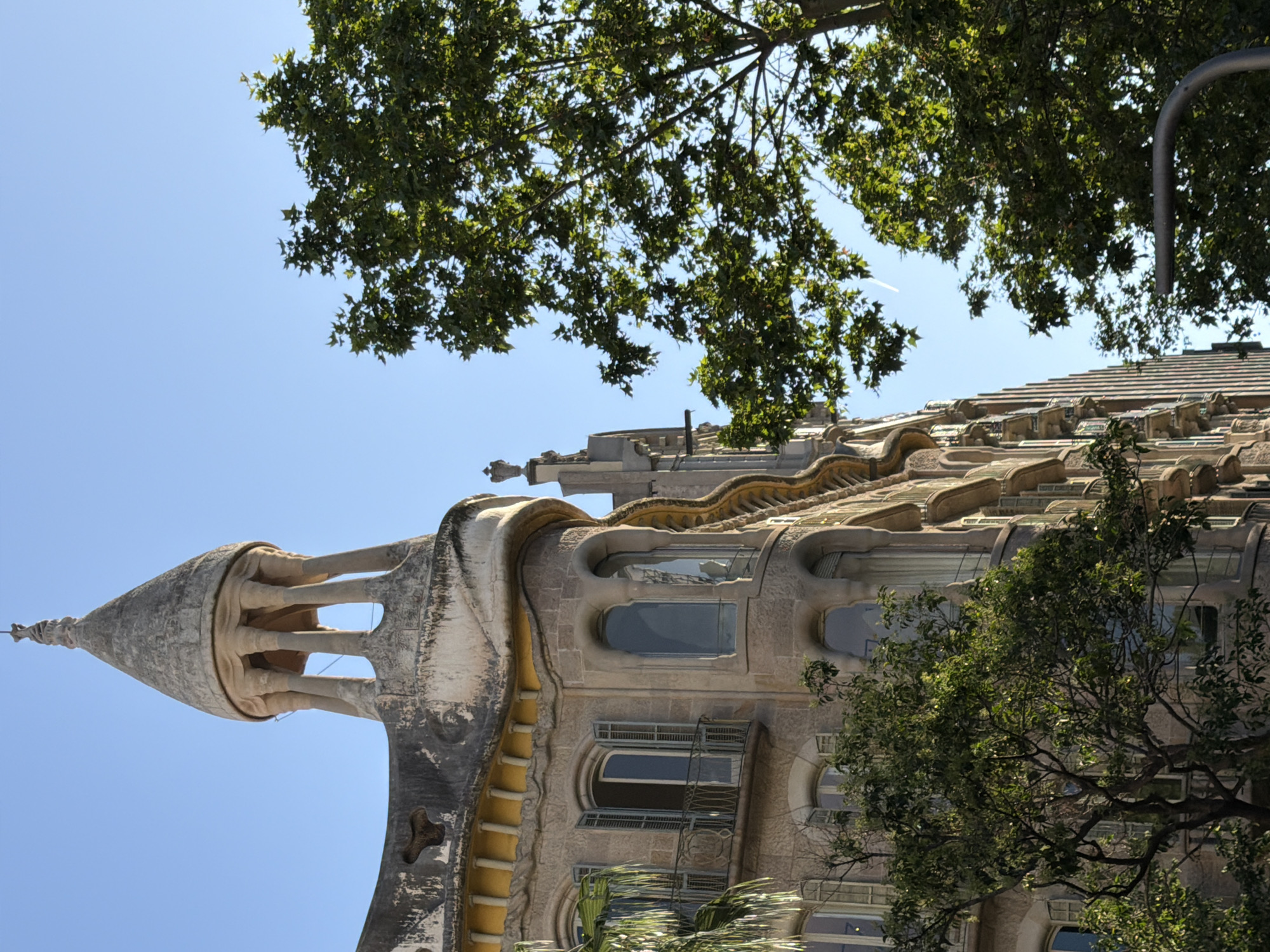 This outdoor photograph captures a unique, organically shaped building, likely in the Modernista architectural style common to Barcelona, Spain. The image is rotated 90 degrees counter-clockwise, causing the building to appear on its side with the sky to the left.
The main subject is a stone or stucco building featuring a prominent conical turret or spire that points to the left. This spire has open, structural elements near its top. The building's facade below is characterized by undulating, wave-like forms and irregularly shaped windows, some deeply recessed and others adorned with small balconies and ornate railings. Decorative, possibly tiled, elements are visible along the curved roofline. The building's color palette primarily consists of warm beige, grey, and some yellowish accents.
Lush green tree foliage partially obscures the building, particularly along what would be its top edge if viewed upright, and on the right side. The background is a clear, bright blue sky, indicating sunny weather during the day. A faint white contrail from an aircraft is visible in the sky. In the upper-right corner, a curved grey metal structure, possibly part of a streetlamp or sign, is partially visible. No people or visible text are present. The perspective is an upward-looking shot, emphasizing the height and unique architectural details of the structure.