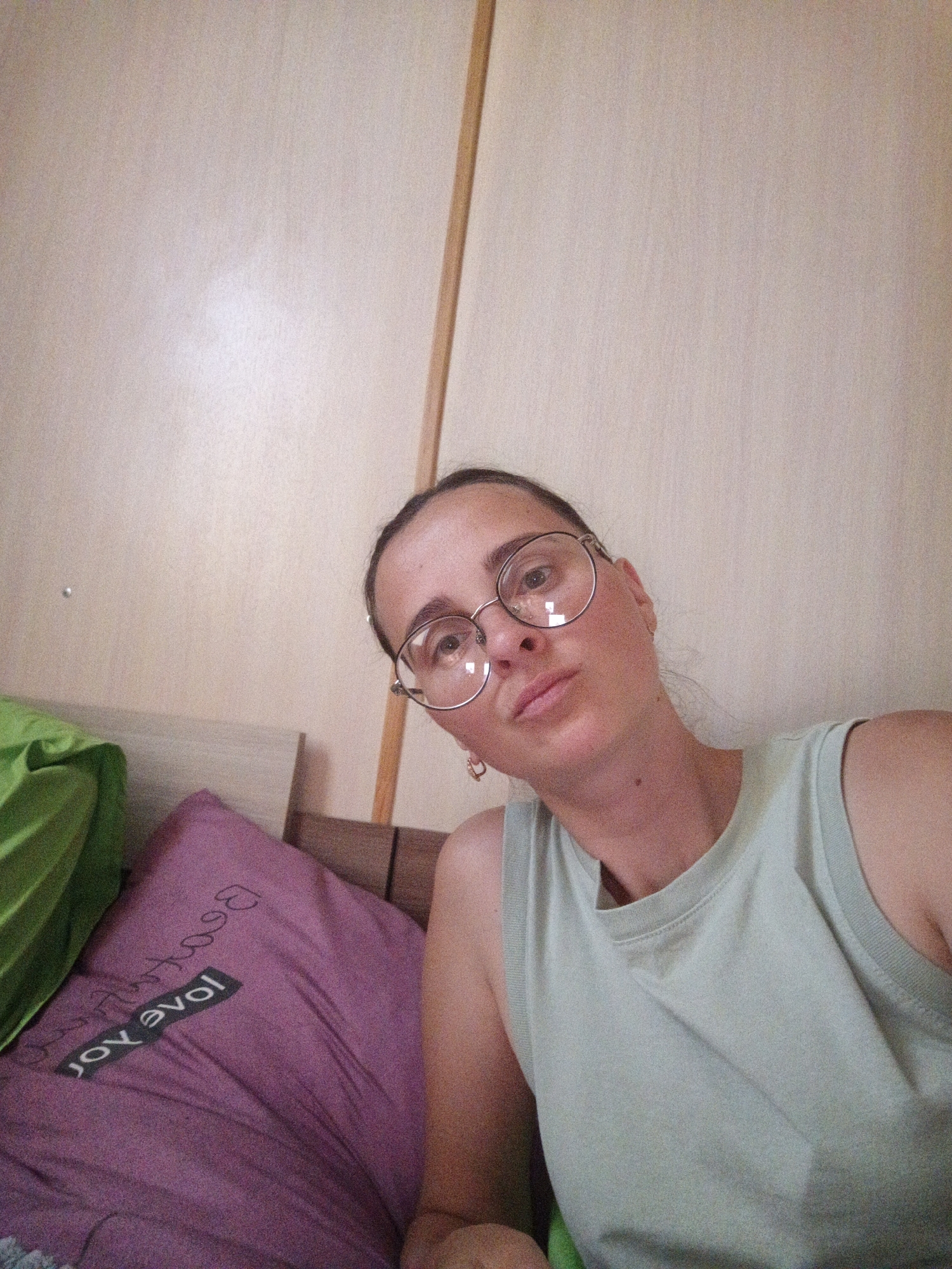 This is a selfie of a woman with brown hair, wearing glasses and a light green tank top. She is positioned on the right side of the frame, leaning towards the camera. Her head is tilted, and she is looking slightly off-camera. She has a neutral expression on her face.
In the background, there is a wall that appears to be made of wood panels. To the left of the woman, a purple pillow is visible with text that reads "love joy" in reverse. A bright green object, possibly a blanket or pillow, is also partially visible on the left. The lighting suggests it might be daytime. The setting appears to be indoors, likely a bedroom given the pillows and the bed. The woman is wearing small hoop earrings. The overall mood is casual and relaxed.
